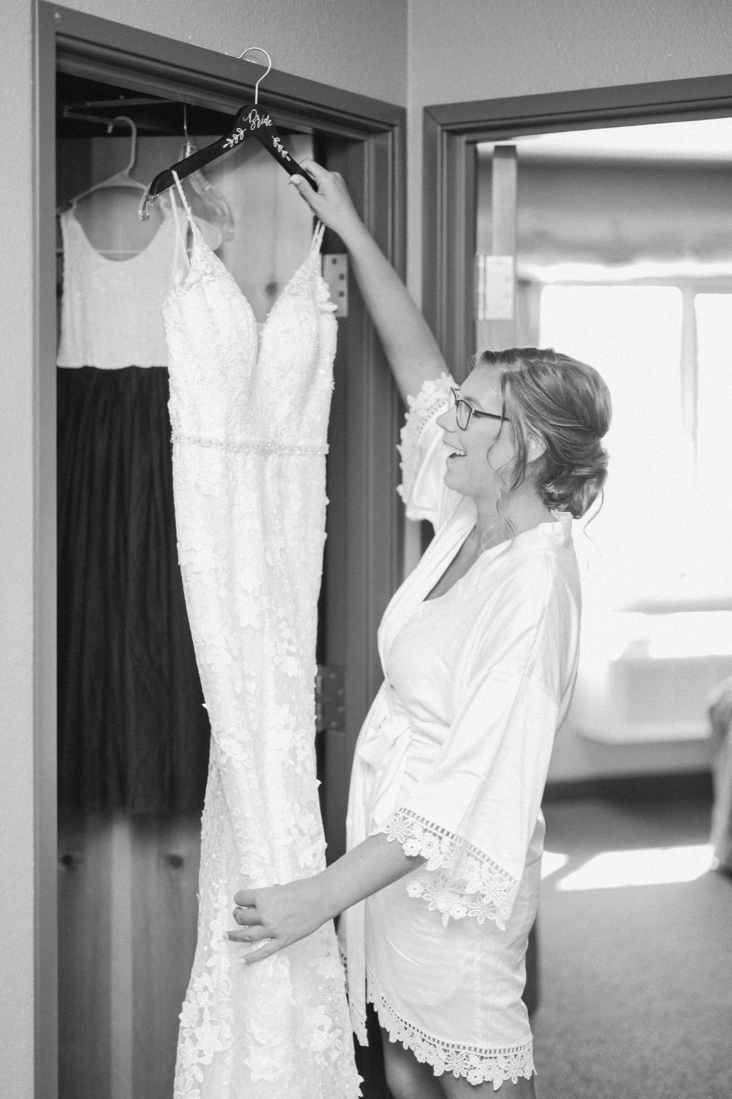 Kim laughs as she reaches up to lift her wedding dress from a closet doorway in a sunlit bridal suite — Tim Larsen Photography, Brainerd Lakes MN