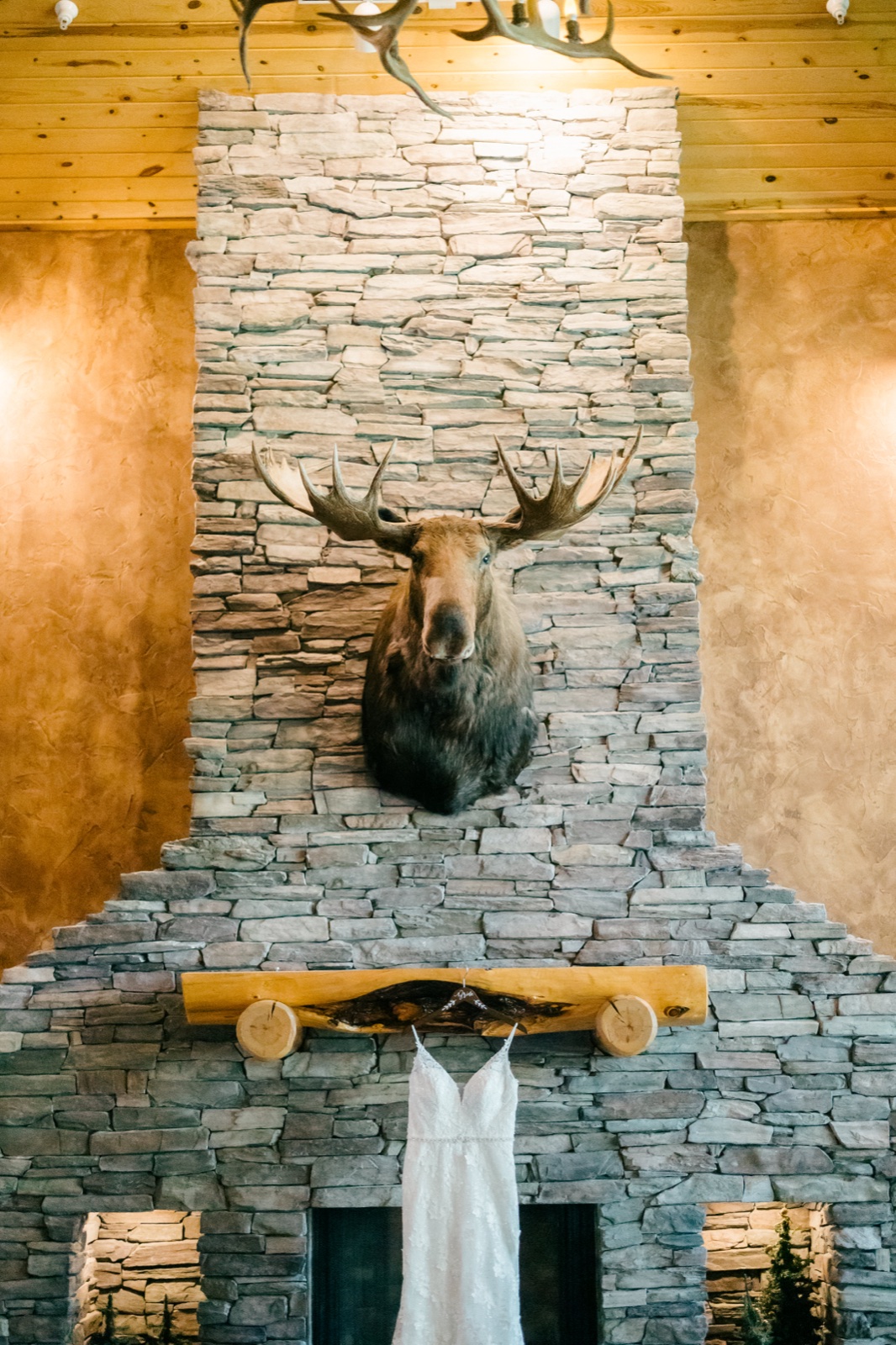 Kim's lace wedding dress hangs from the wooden mantel of a tall stone fireplace beneath a mounted moose head and antler chandelier in the Whitefish Lodge great room — Tim Larsen Photography, Brainerd Lakes MN
