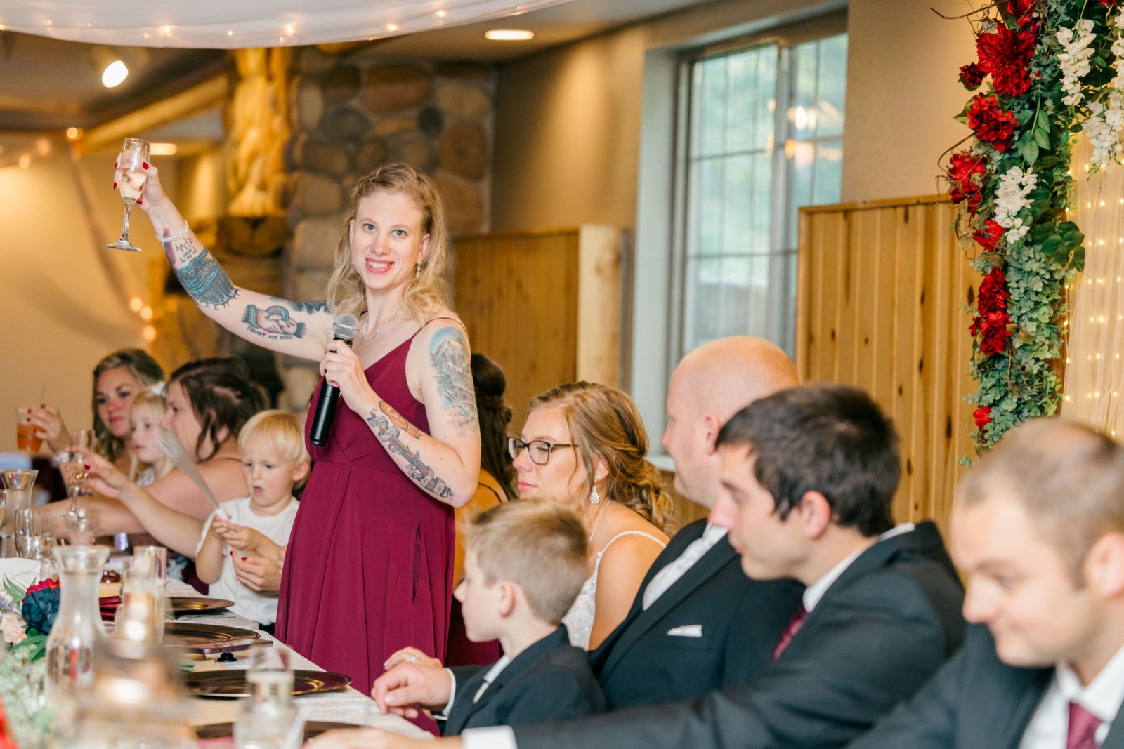 A bridesmaid in a burgundy dress raises a champagne glass while holding a microphone during a toast at the reception head table — Tim Larsen Photography, Brainerd Lakes MN