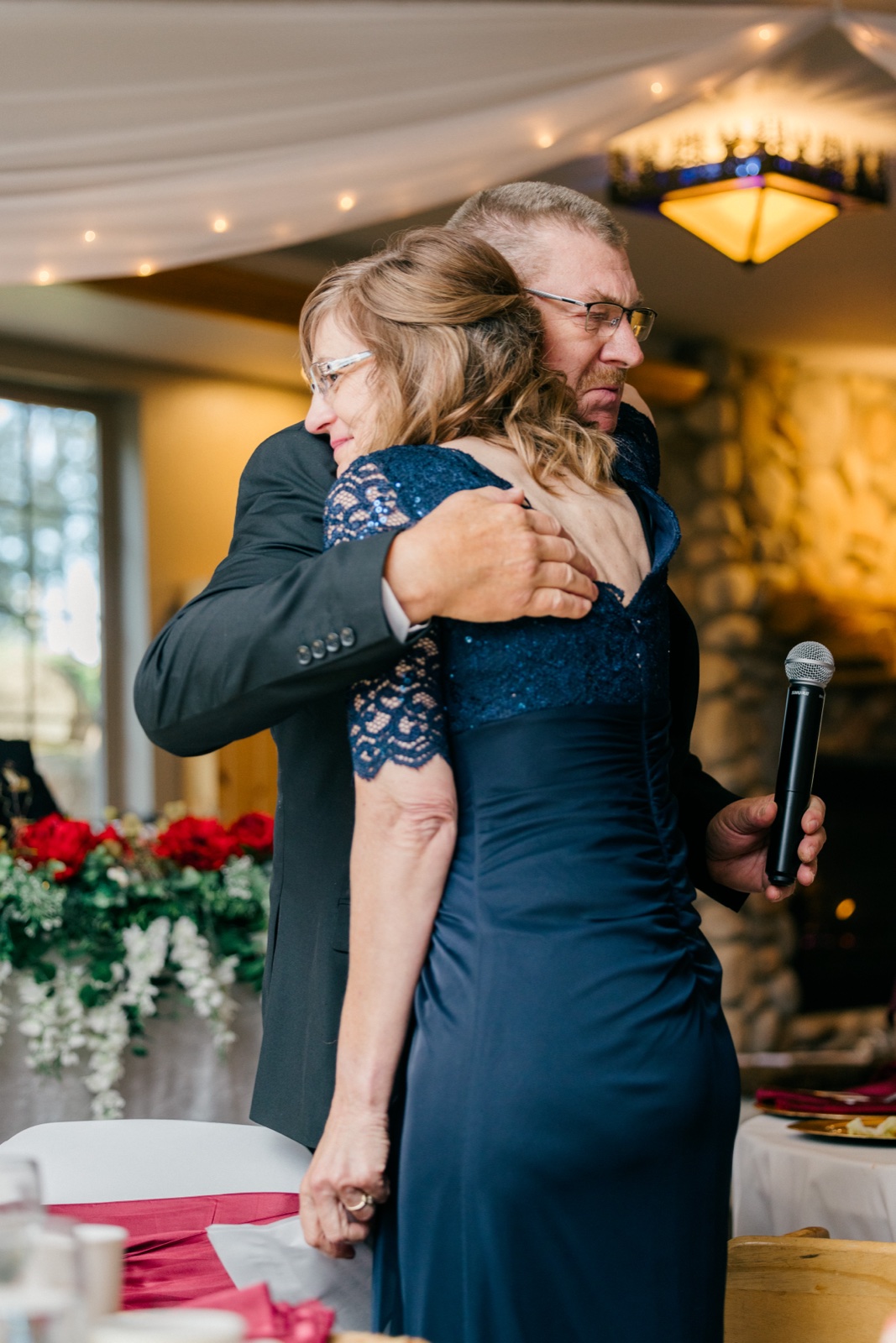 A father and daughter share a quiet embrace after a toast, the father holding a microphone behind her back as she rests her cheek against his shoulder — Tim Larsen Photography, Brainerd Lakes MN