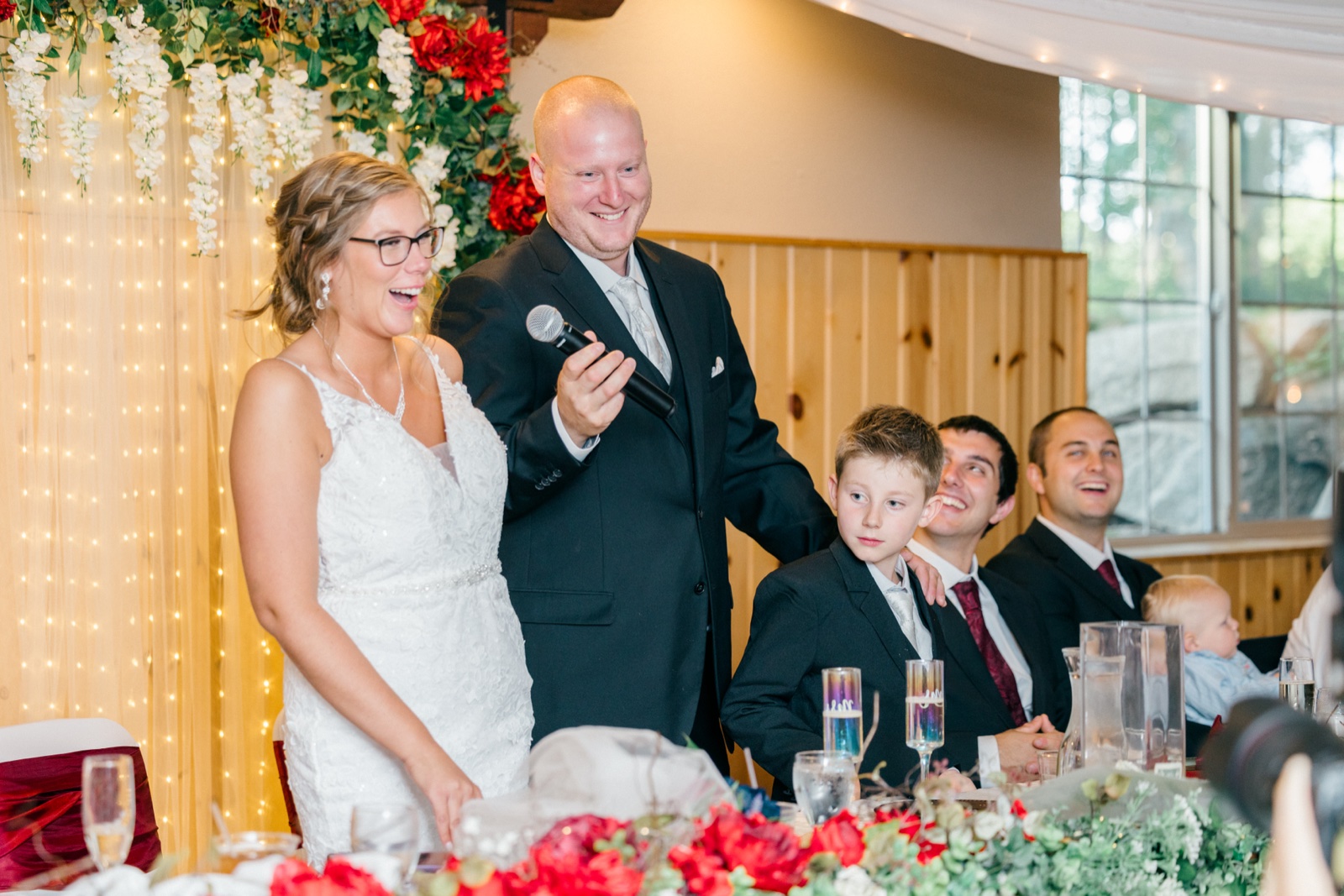 Kim laughs while Jesse holds a microphone giving a toast, with groomsmen and a young ring bearer reacting with smiles at the head table — Tim Larsen Photography, Brainerd Lakes MN