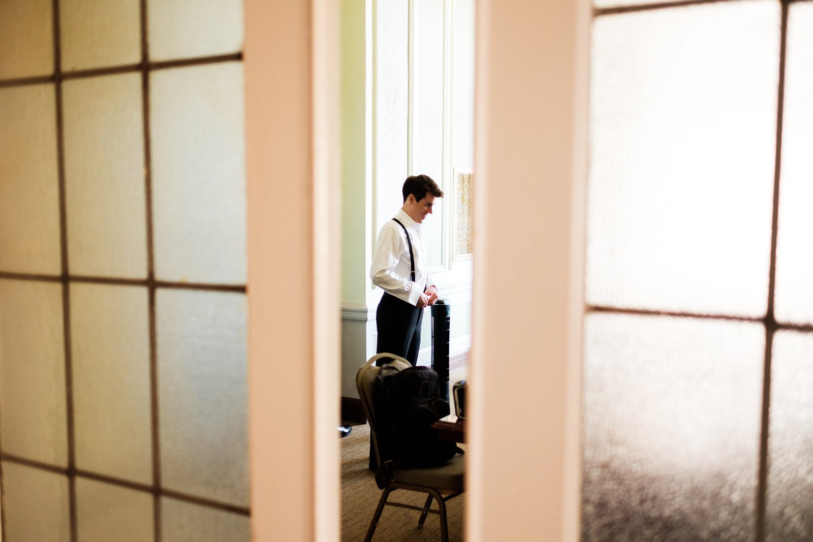 The groom stands in window light buttoning his cuff, glimpsed through a partially open frosted glass door — Tim Larsen Photography, Duluth MN