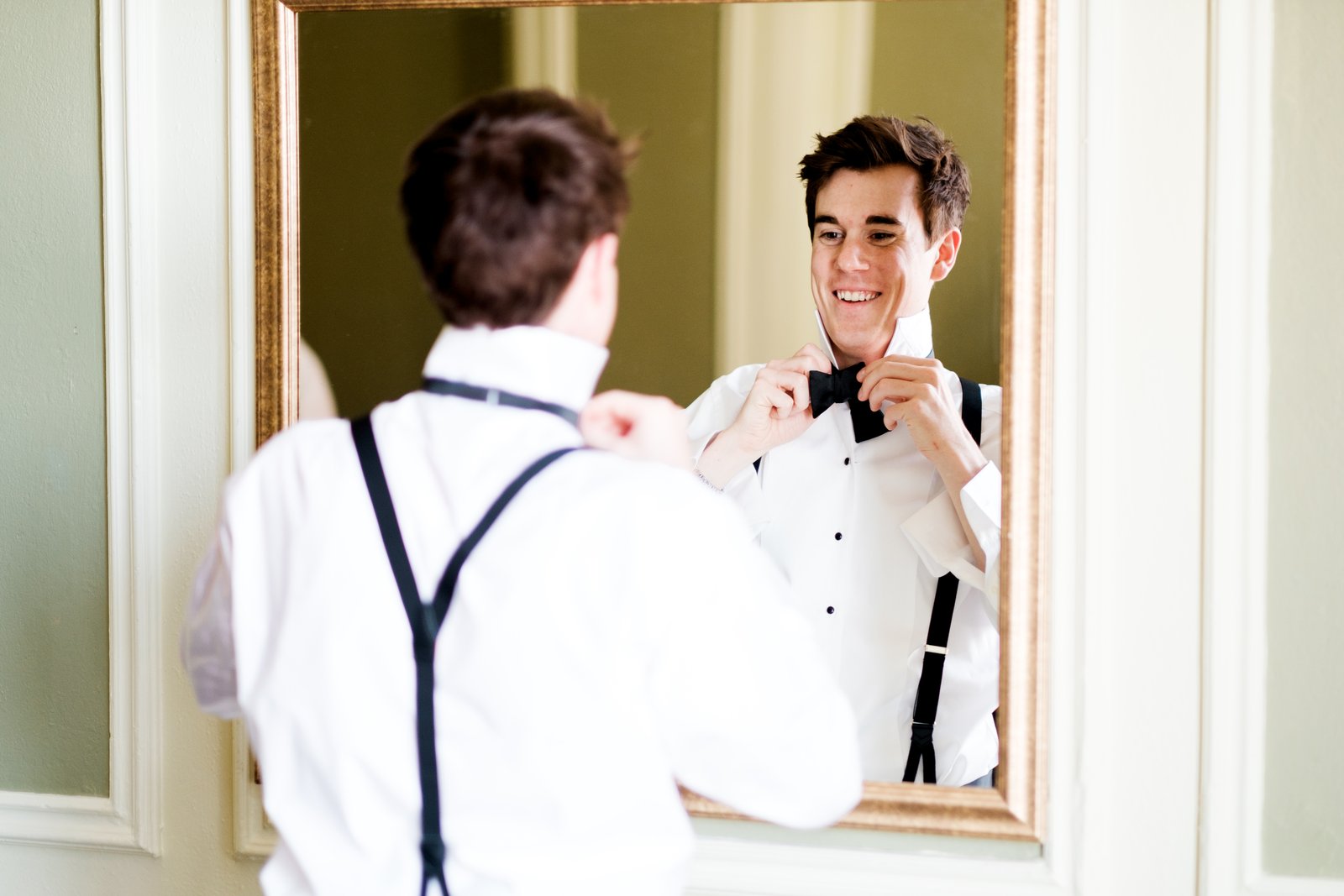 The groom smiles broadly at his own reflection in an ornate gilded mirror as he ties his black bow tie, suspendered back to the camera — Tim Larsen Photography, Duluth MN