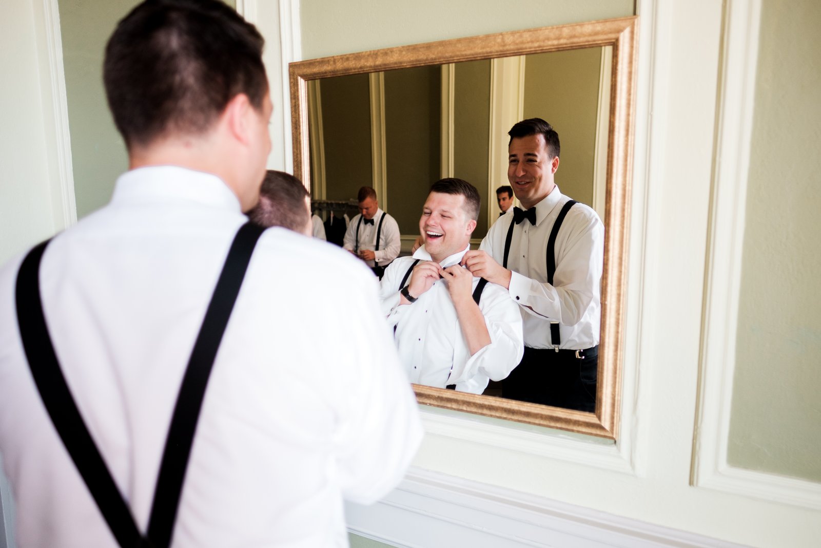 A groomsman in suspenders watches in the foreground as the groom laughs while a friend ties his bow tie, reflected in a large framed mirror — Tim Larsen Photography, Duluth MN