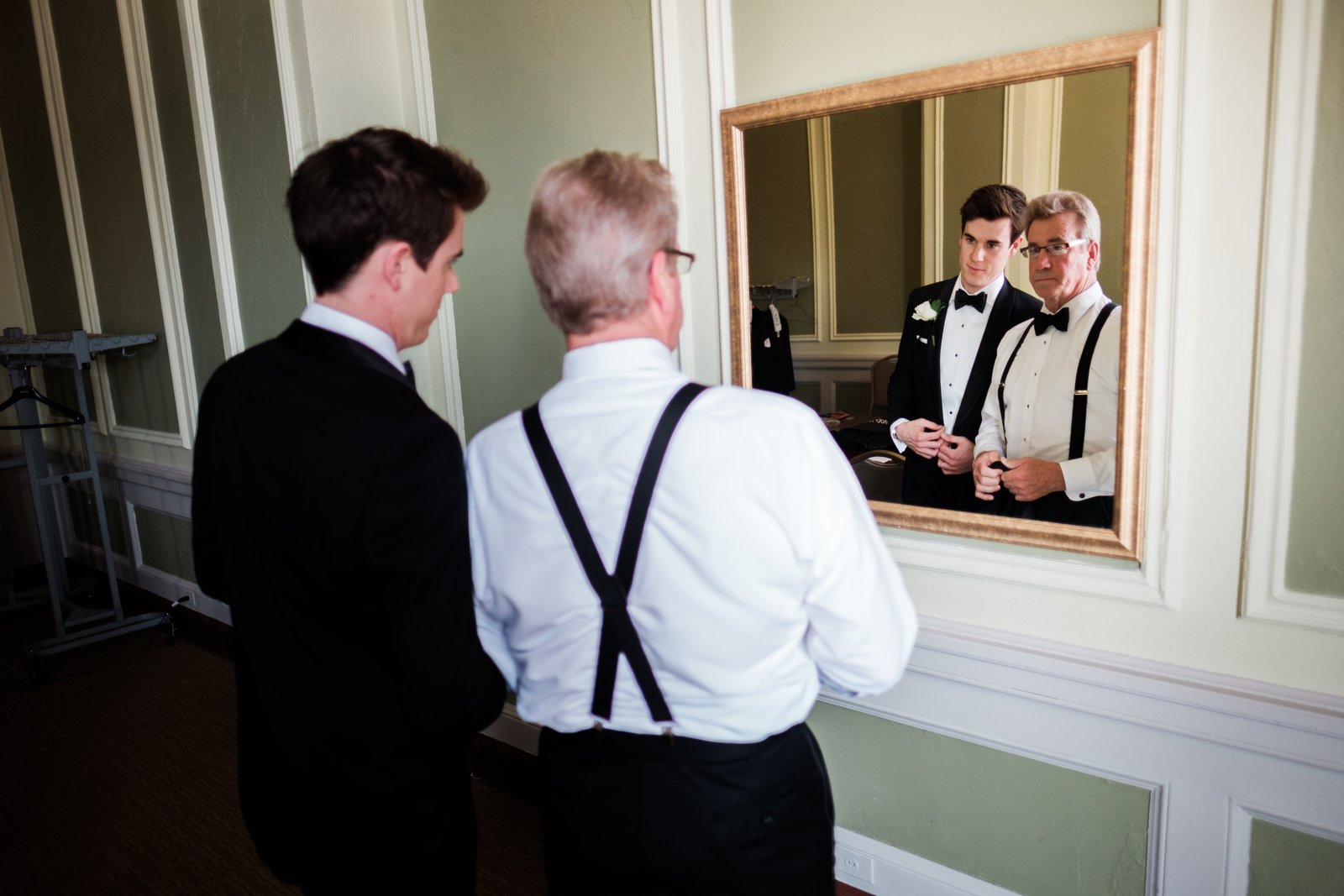 The groom and his father stand together adjusting bow ties, their reflection in a large gilded mirror while a younger groomsman watches from the left — Tim Larsen Photography, Duluth MN