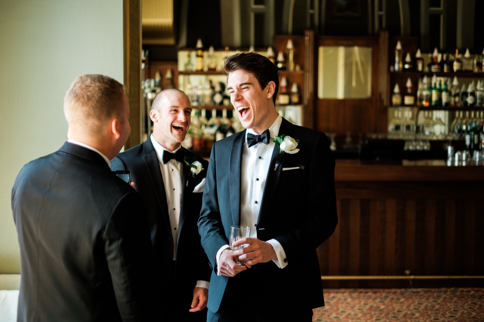 The groom laughs open-mouthed beside two groomsmen at an ornate wood bar during cocktail hour inside the Greysolon Plaza — Tim Larsen Photography, Duluth MN
