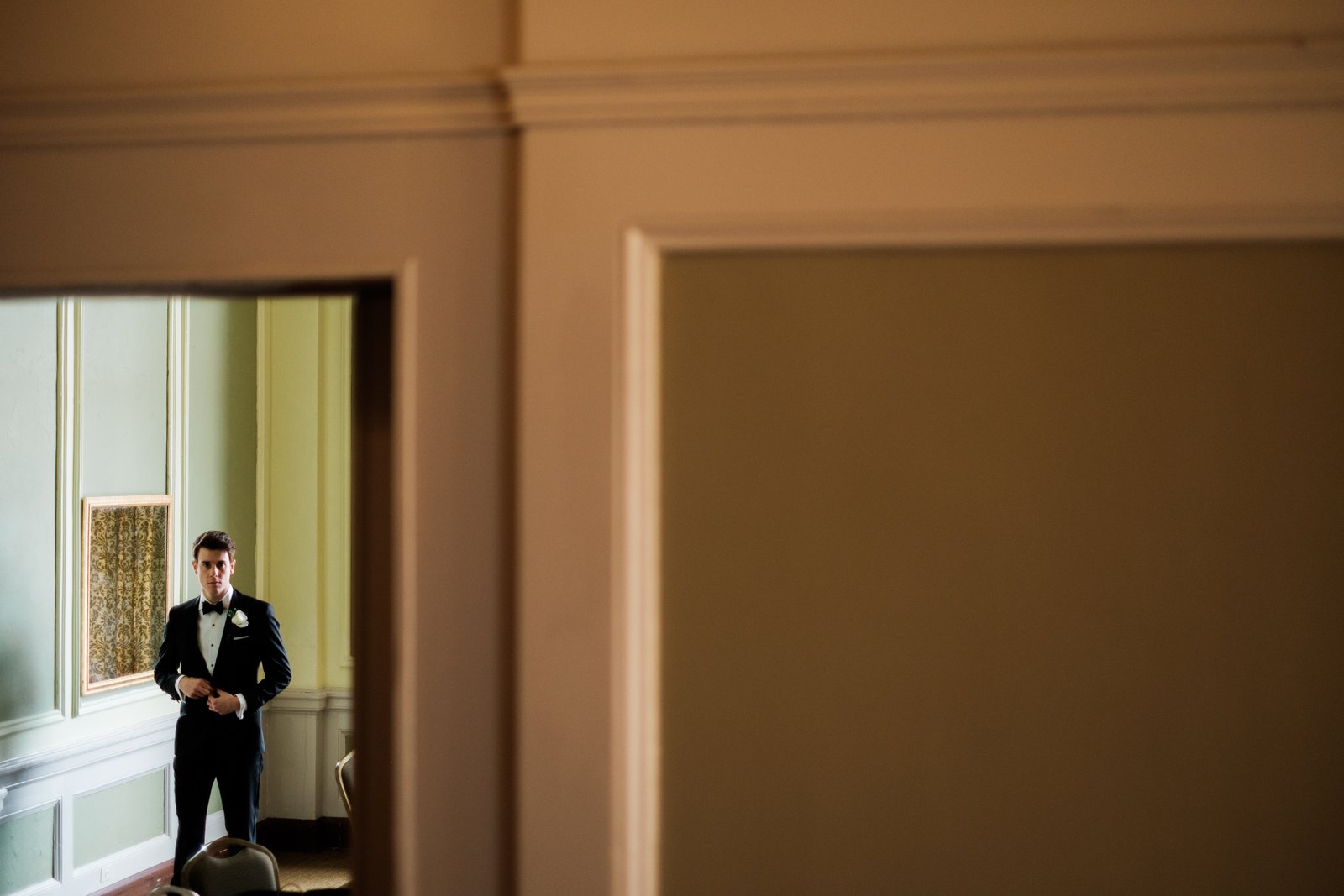 The groom in a black tuxedo buttons his jacket in window light, seen through a distant doorway with a large warm-toned wall dominating the right side of the frame — Tim Larsen Photography, Duluth MN