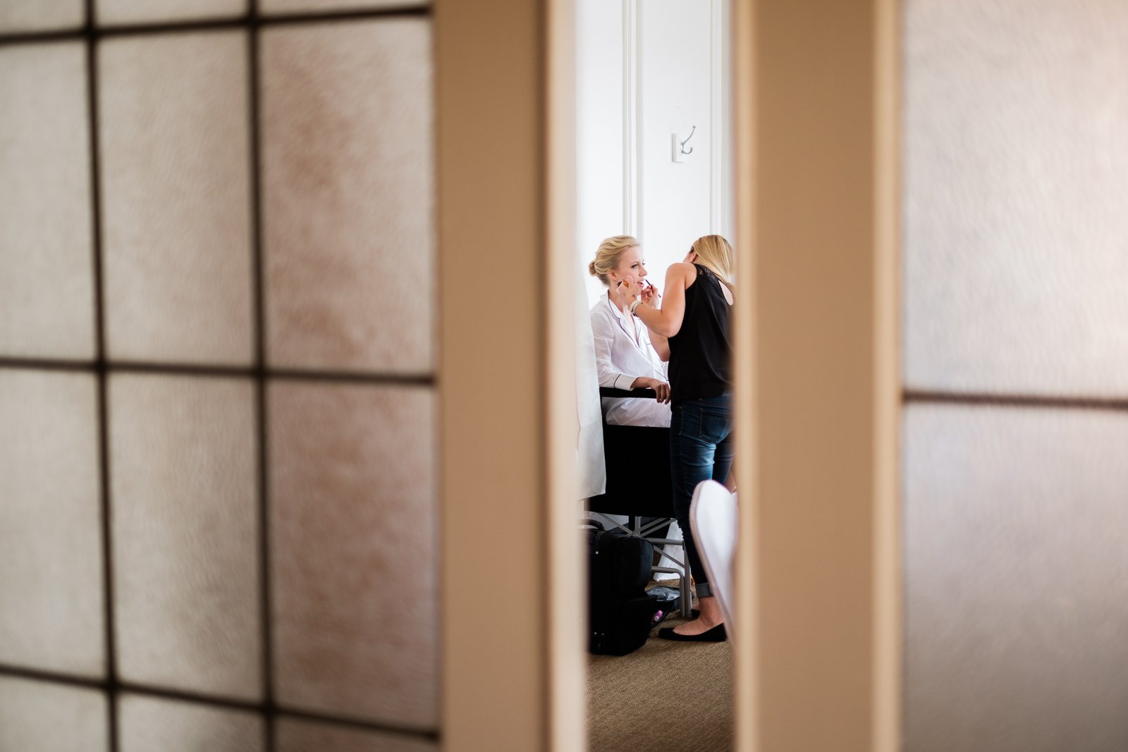 The bride sits in window light having her makeup applied, viewed through the narrow opening of a shoji-style sliding door — Greysolon Plaza bridal suite — Tim Larsen Photography, Duluth MN