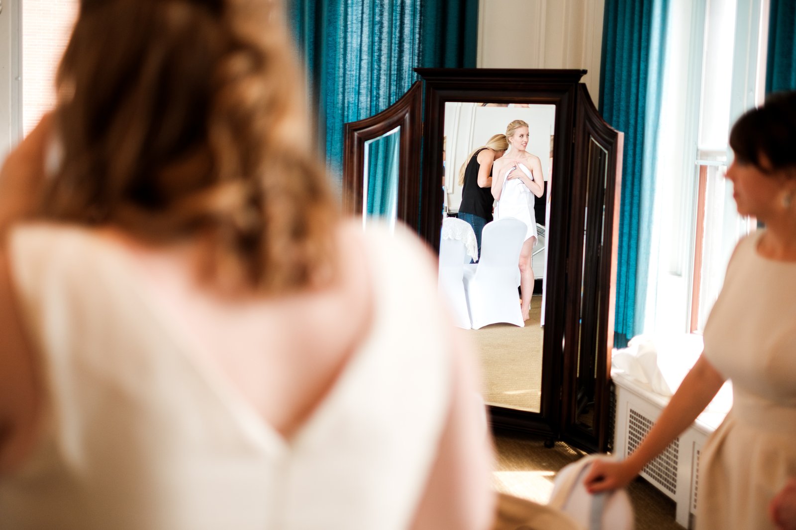 The bride reflected in a tall mirror as her mother fastens her dress, watched by two bridesmaids in the soft-focus foreground — Tim Larsen Photography, Duluth MN