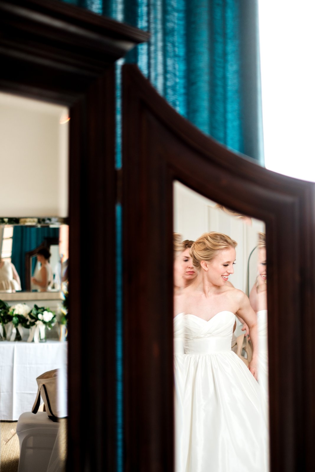 The bride smiles softly while dressing, her reflection layered through an ornate wardrobe mirror and doorway with rich teal curtains framing the suite — Tim Larsen Photography, Duluth MN