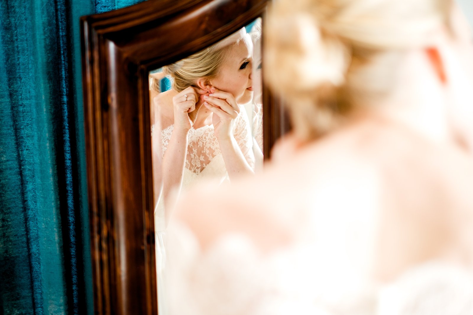 The bride fastens an earring, framed in a carved wooden mirror over her own out-of-focus shoulder against teal velvet drapery — Tim Larsen Photography, Duluth MN