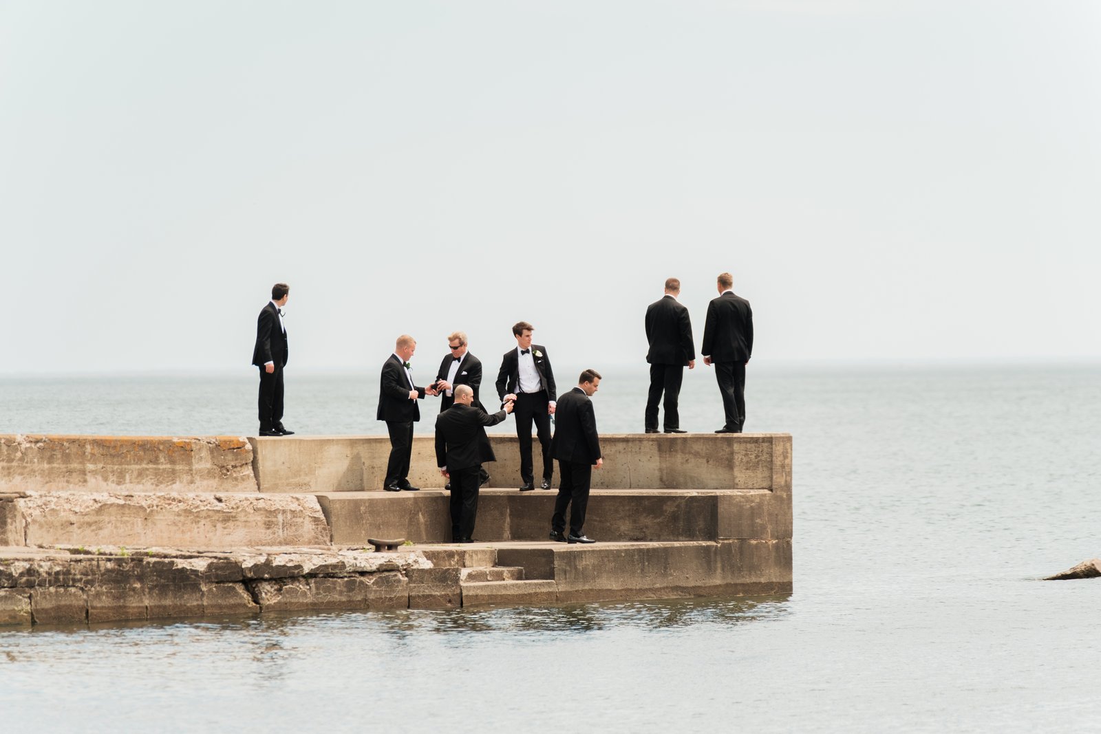 Eight groomsmen in black tuxedos stand scattered across a concrete pier extending into Lake Superior under an overcast sky — Tim Larsen Photography, Duluth MN