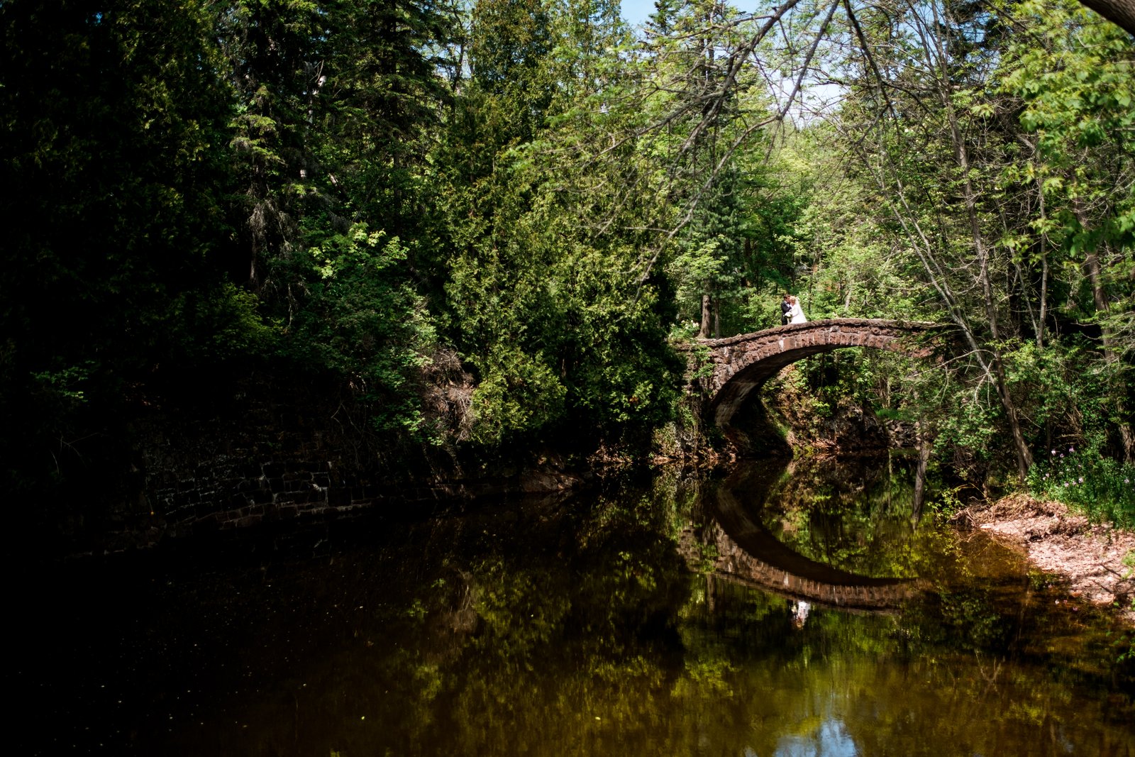 The bride and groom embrace on a stone arch bridge over a still creek, surrounded by dense green forest with their reflection visible in the water below — Tim Larsen Photography, Duluth MN