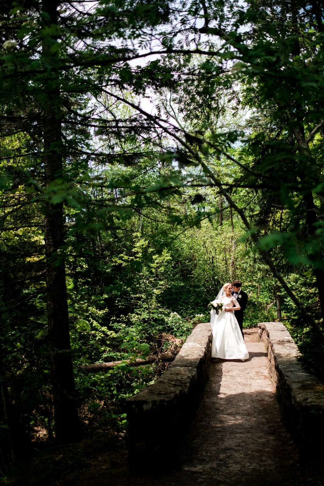 The bride and groom embrace on a sunlit stone footbridge, framed by an arching tunnel of dense green forest foliage — Tim Larsen Photography, Duluth MN