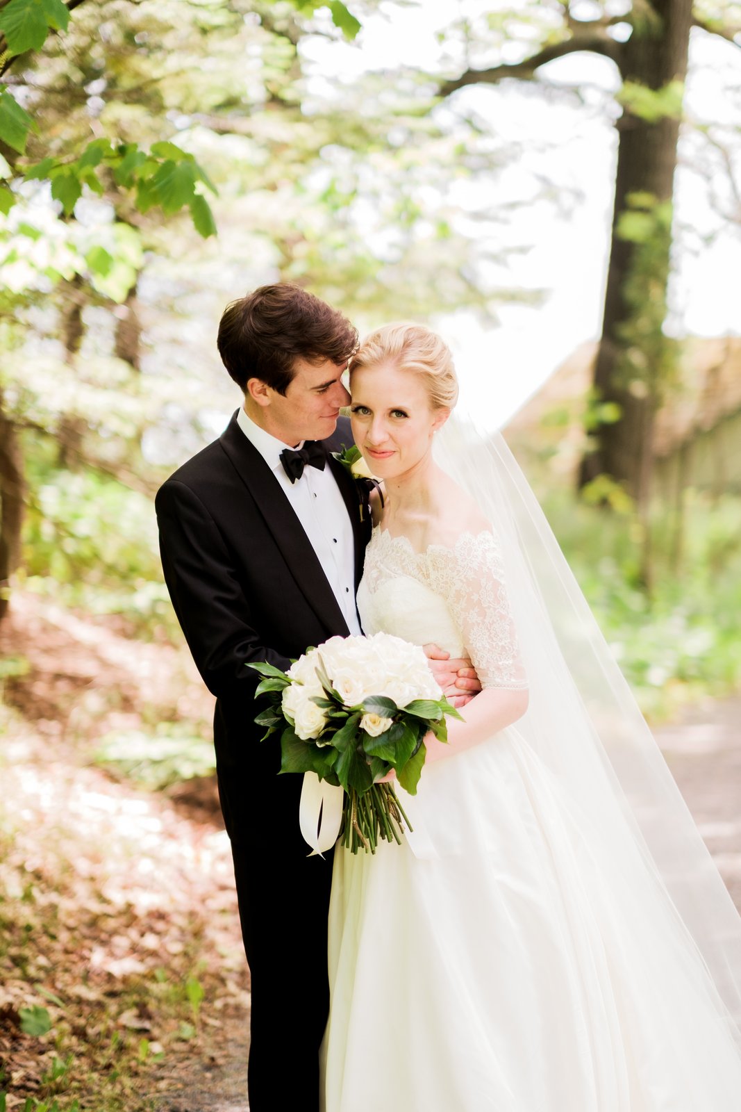 The groom leans into the bride's temple as she looks calmly to the camera, her veil trailing into the sunlit trees behind them — Tim Larsen Photography, Duluth MN