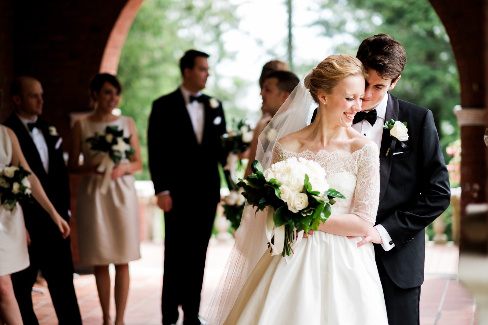 The groom leans into the bride's cheek as she laughs holding her white rose bouquet, the bridal party softly visible behind them beneath a brick archway — Tim Larsen Photography, Duluth MN