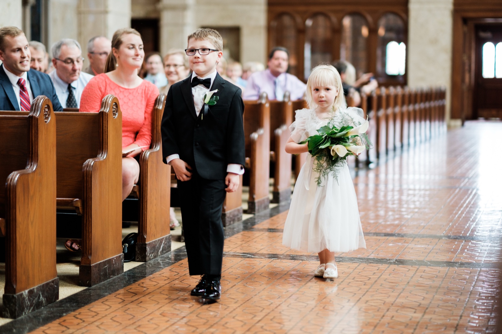 A young ring bearer in a black tuxedo and a flower girl in a white dress holding a bouquet walk down the cathedral aisle past seated guests in wooden pews — Tim Larsen Photography, Duluth MN