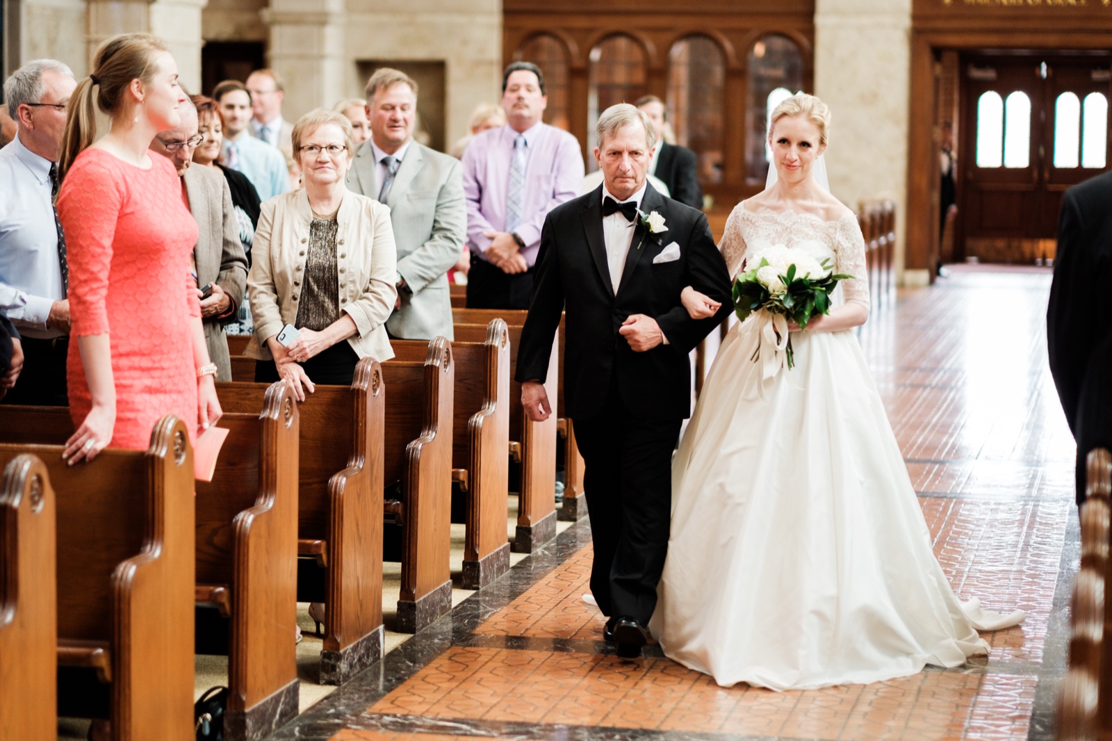 The bride walks the long center aisle on her father's arm in a lace-sleeve ball gown and cathedral veil as guests stand watching from the wooden pews — Tim Larsen Photography, Duluth MN