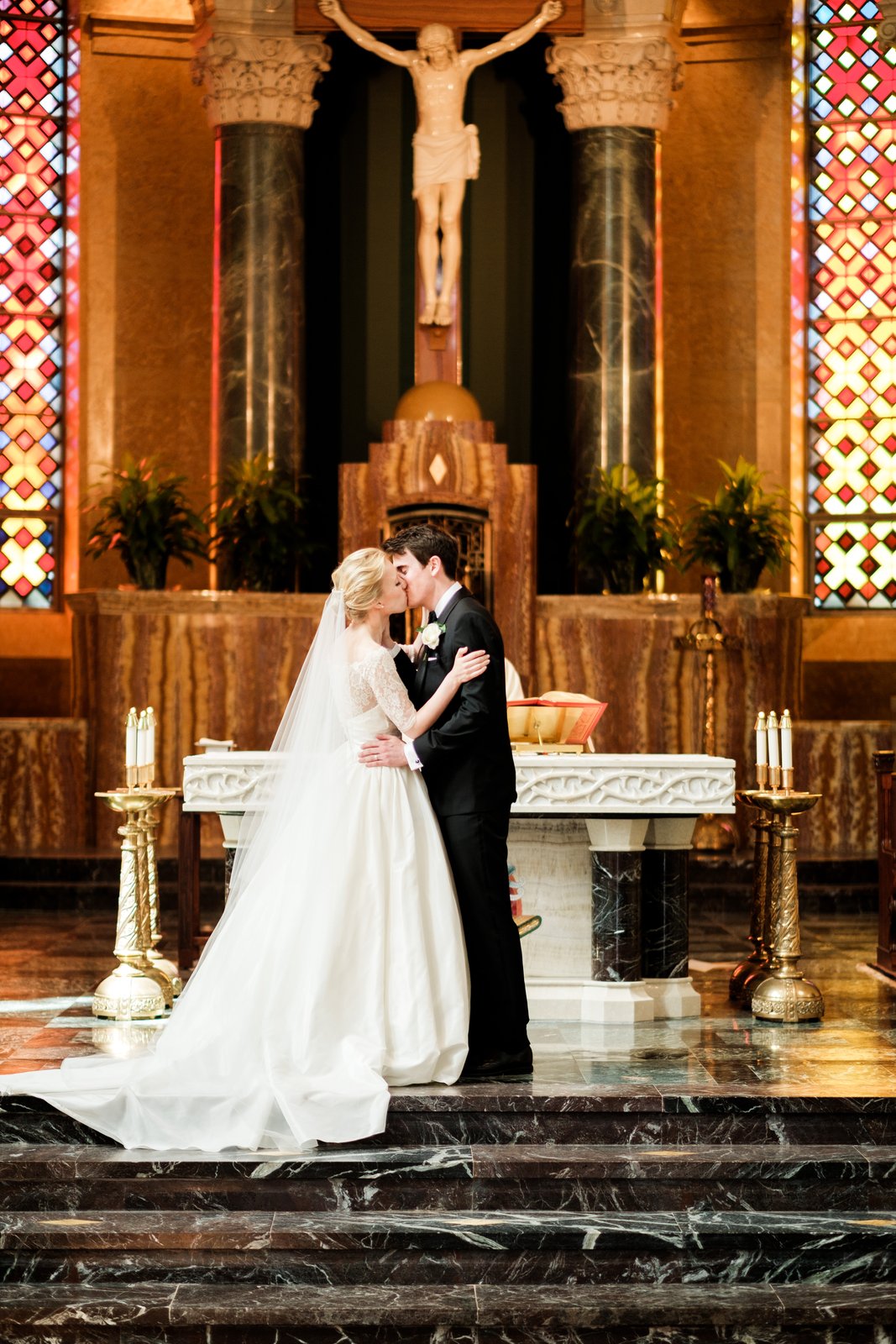 The bride and groom kiss at the altar beneath a crucifix flanked by stained glass windows — Cathedral of Our Lady of the Rosary, Duluth — Tim Larsen Photography, Duluth MN