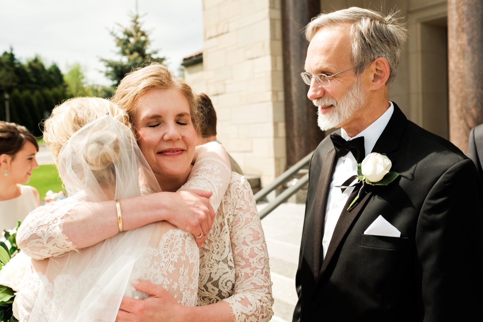 The bride embraces her mother on the Cathedral steps with her eyes closed while her father watches with a soft smile from the right — Tim Larsen Photography, Duluth MN
