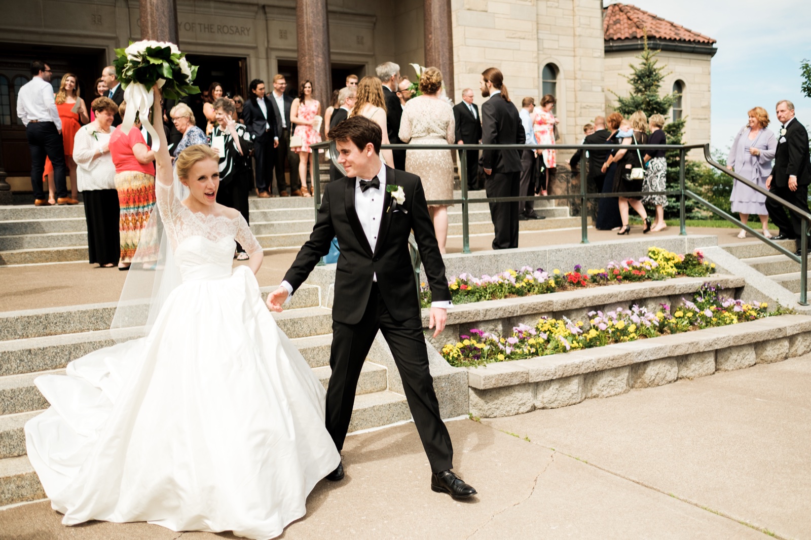 The bride raises her bouquet overhead in triumph as the groom leads her down the stone Cathedral steps past rows of cheering guests and beds of bright flowers — Tim Larsen Photography, Duluth MN