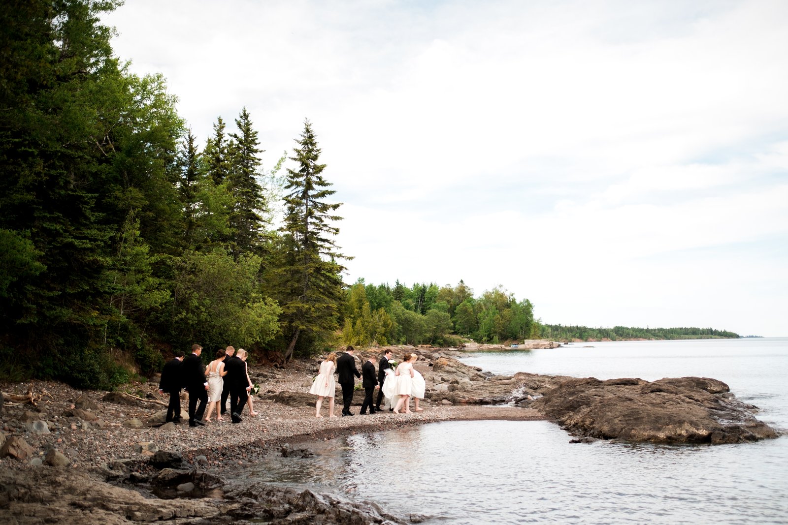The small bridal party walks along a rocky Lake Superior shoreline beneath tall evergreens, the open lake stretching to the horizon — Tim Larsen Photography, Duluth MN