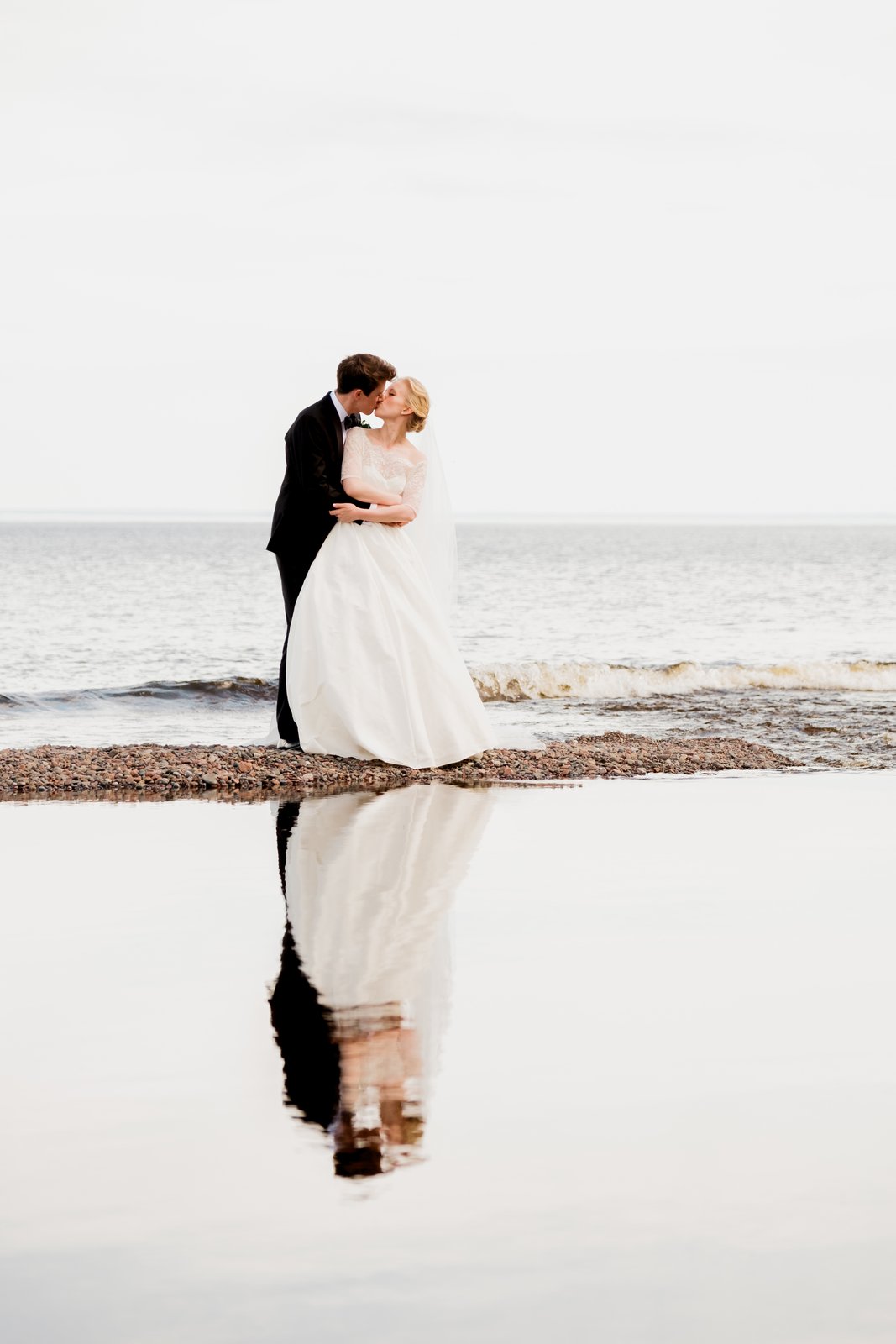 The bride and groom kiss on the rocky Lake Superior shore, their full figures perfectly mirrored in a still tidal pool in the foreground — Tim Larsen Photography, Duluth MN