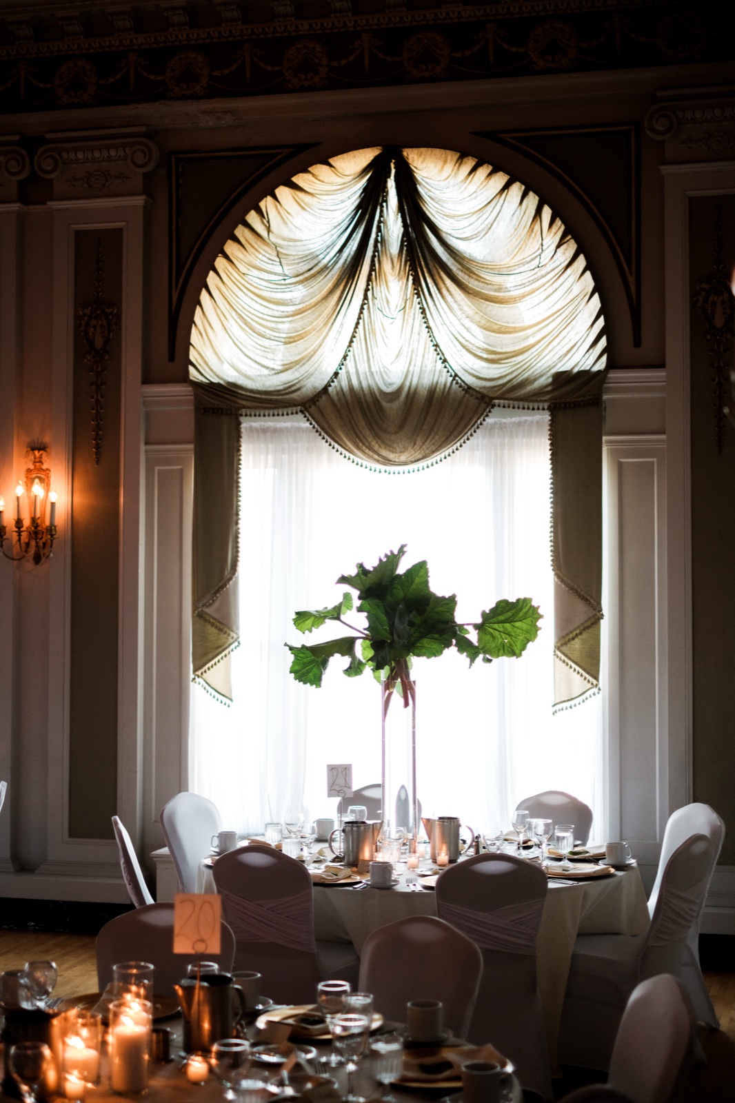 A tall rhubarb-leaf centerpiece rises in front of a grand draped arched window in the Greysolon Ballroom above set reception tables and chair-covered seats — Tim Larsen Photography, Duluth MN