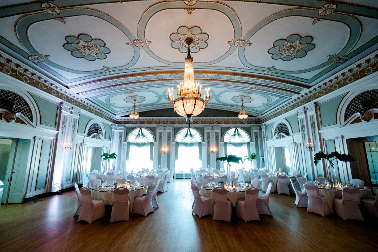 The historic Greysolon Ballroom set for a wedding dinner — a grand crystal chandelier hanging from the frescoed ceiling above round chair-covered reception tables — Tim Larsen Photography, Duluth MN