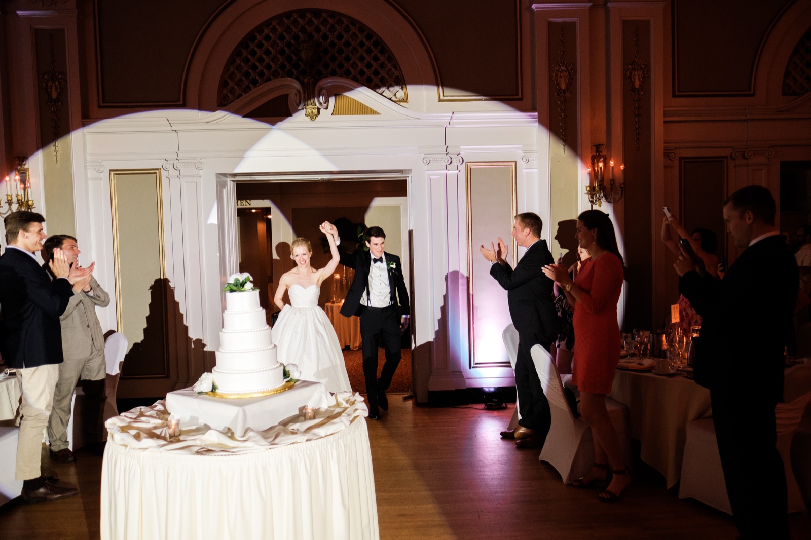 The bride raises her arm in triumph as she and the groom enter the Greysolon Ballroom through an ornate doorway, guests applauding as a tall tiered cake stands in the foreground — Tim Larsen Photography, Duluth MN