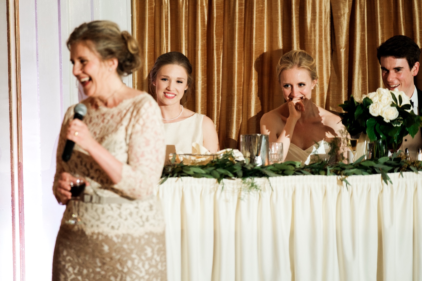 The bride covers her mouth laughing while seated at the head table as her mother delivers a toast in the soft-focus foreground — Tim Larsen Photography, Duluth MN