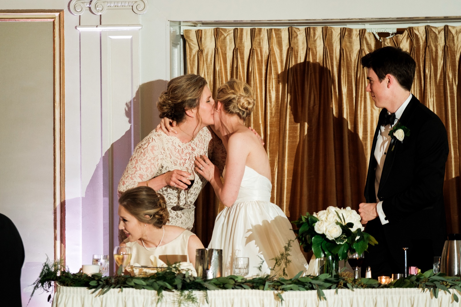 The bride leans across the head table to kiss her mother on the cheek while the groom looks on from the right — Tim Larsen Photography, Duluth MN