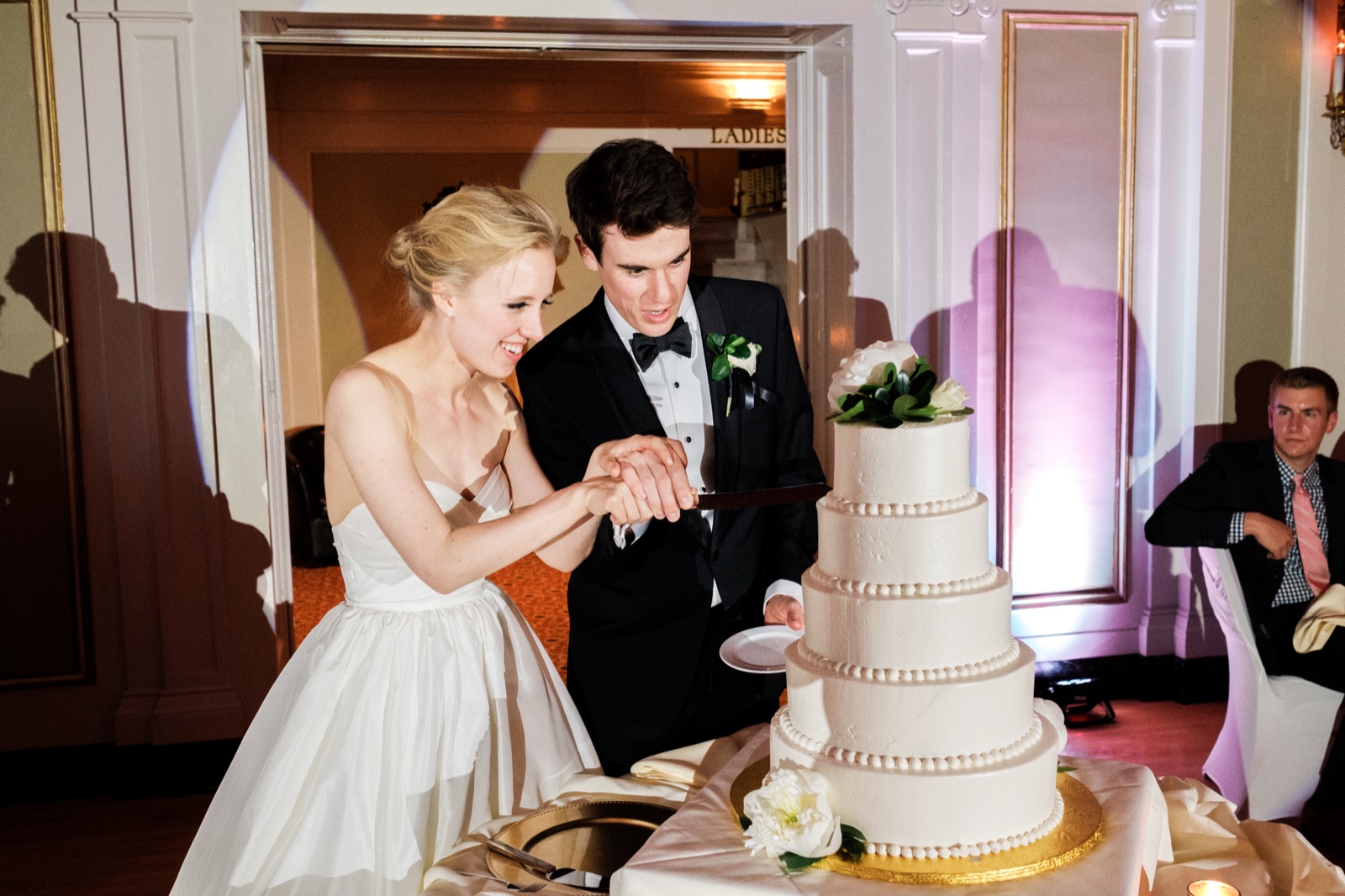 The bride and groom grip a knife together and lean in to cut a tall white tiered wedding cake, their shadows cast on the wall behind them — Tim Larsen Photography, Duluth MN