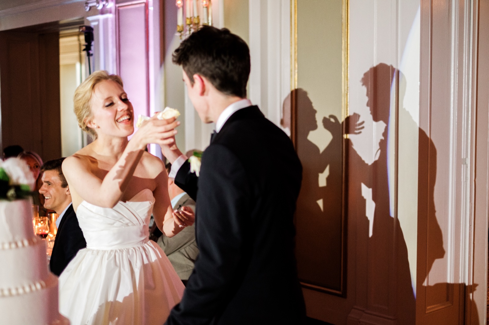 The bride laughs as she feeds the groom a bite of cake, their silhouettes echoing the moment on the wall behind them — Greysolon Ballroom reception — Tim Larsen Photography, Duluth MN