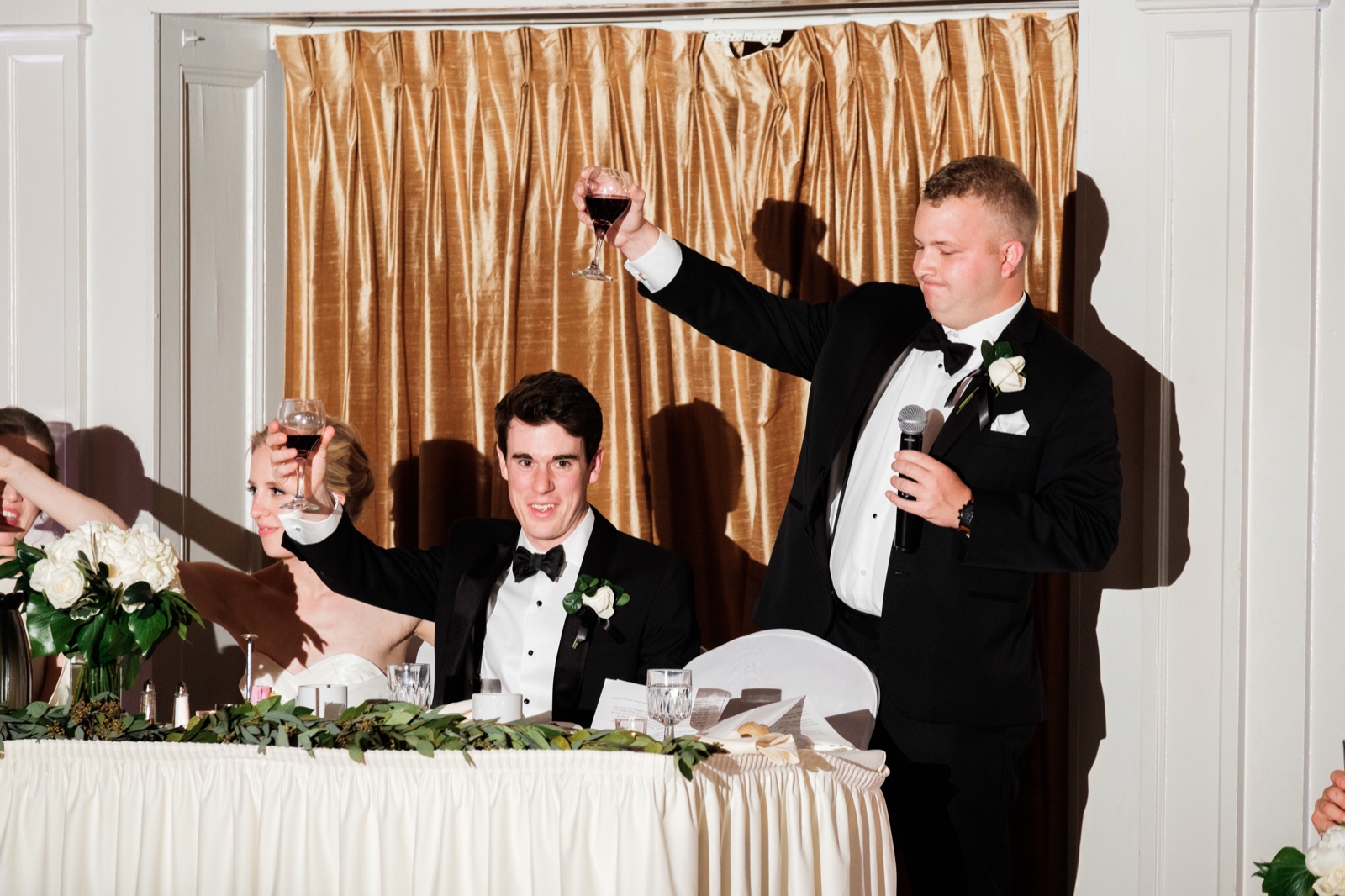 The best man raises a glass and microphone during a toast as the groom reacts and the bride lifts her glass behind him at the head table — Tim Larsen Photography, Duluth MN