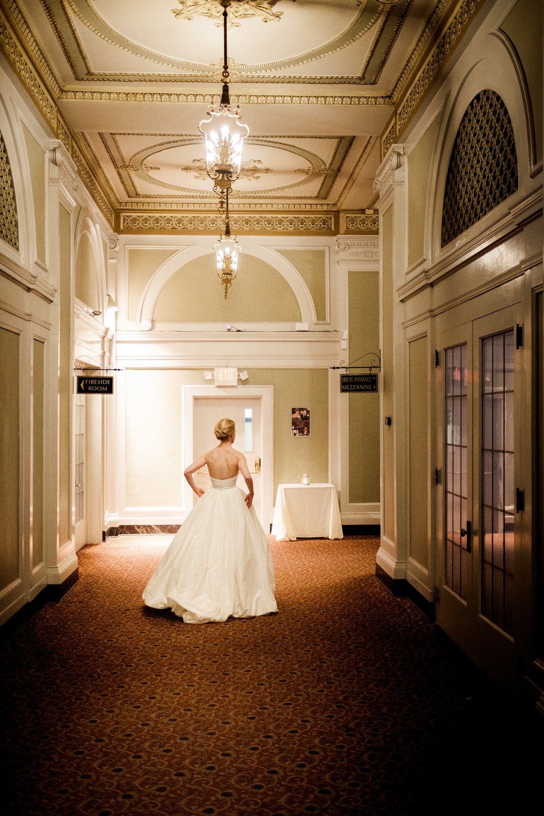 The bride stands alone in a grand historic Greysolon Plaza hallway beneath ornate ceiling medallions and hanging lanterns, her ball gown spreading across the patterned carpet — Tim Larsen Photography, Duluth MN