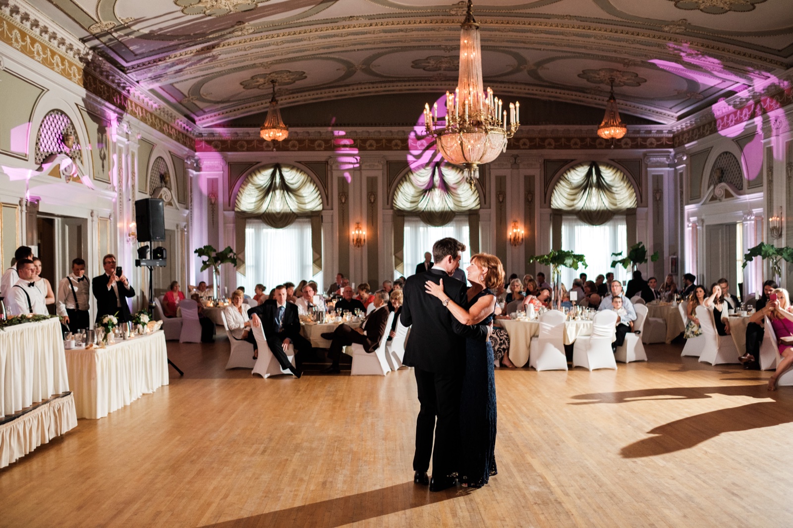 The groom dances with his mother on the empty ballroom floor beneath an ornate chandelier as seated guests watch from tables ringing the historic Greysolon Ballroom — Tim Larsen Photography, Duluth MN
