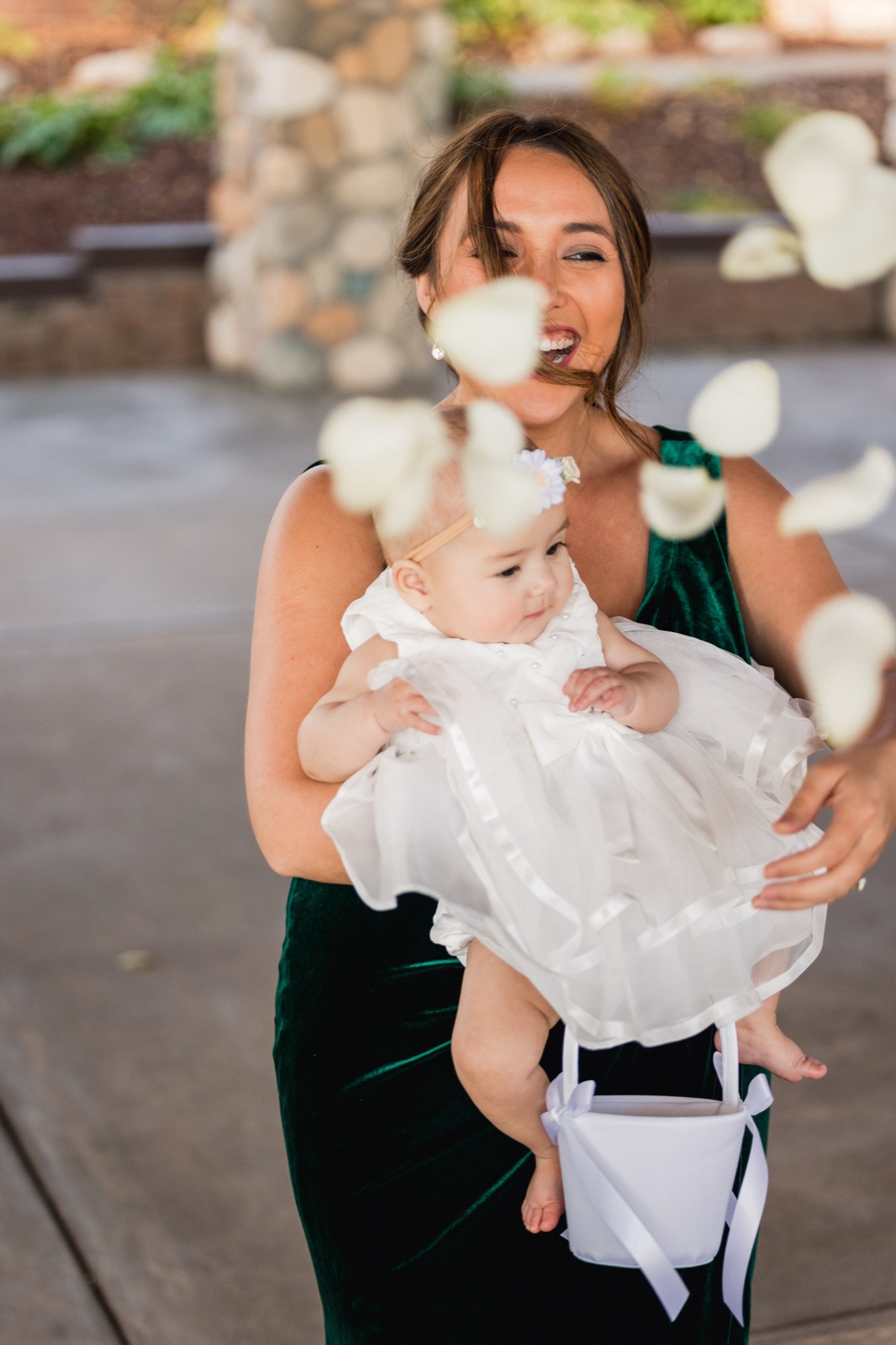 A woman in a green velvet dress laughs while holding a baby flower girl in a white dress as petals fly through the air around them — Tim Larsen Photography, Brainerd Lakes MN