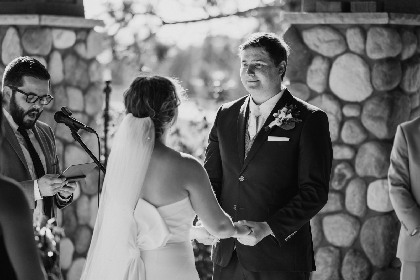 In black and white, the groom holds the bride's hands and gazes at her during the ceremony while the officiant reads from notes beside a stone pillar — Tim Larsen Photography, Brainerd Lakes MN