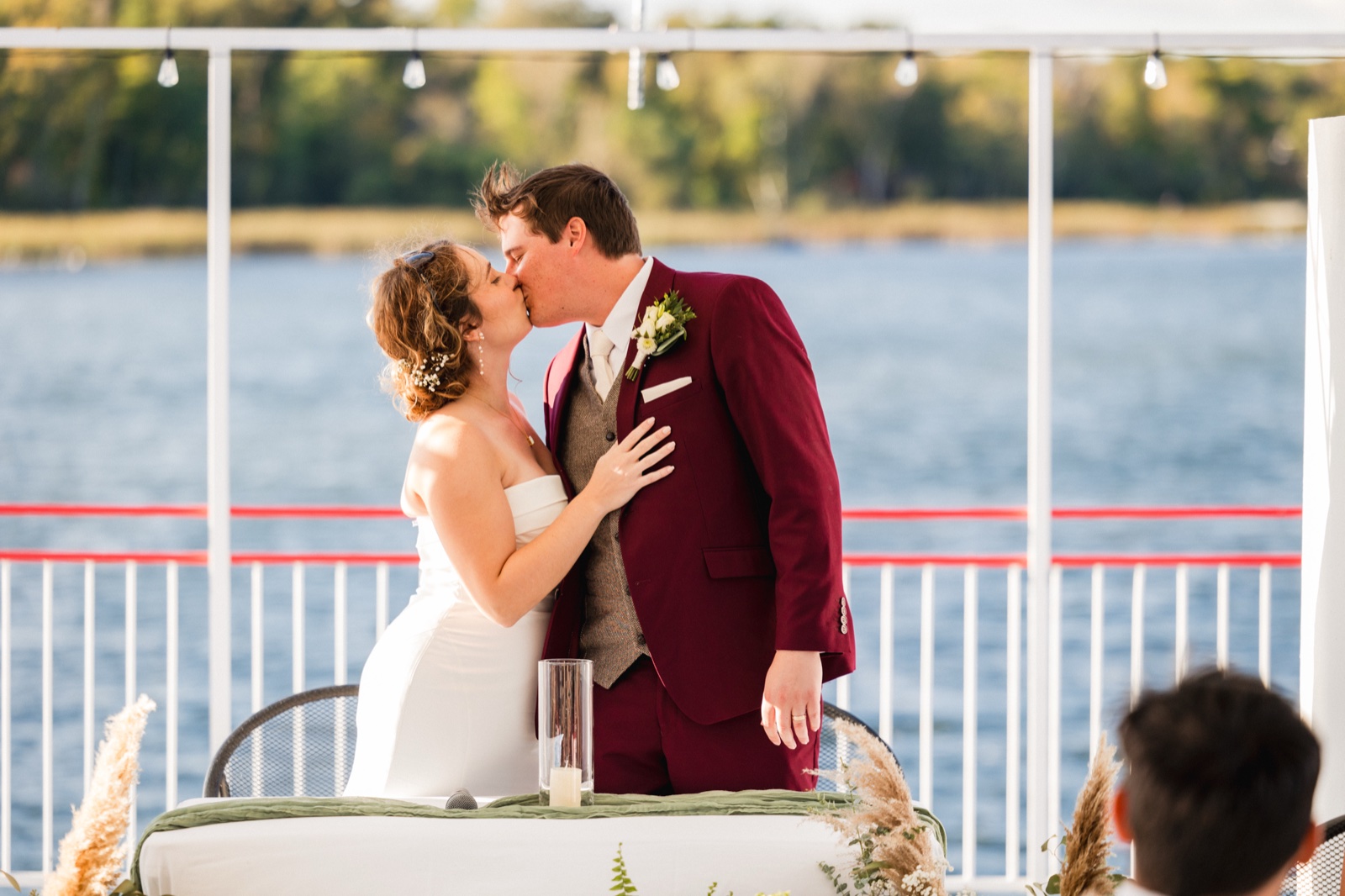 The bride and groom share their first kiss at a lakeside ceremony, with open water and a string-lit pergola behind them — Tim Larsen Photography, Brainerd Lakes MN