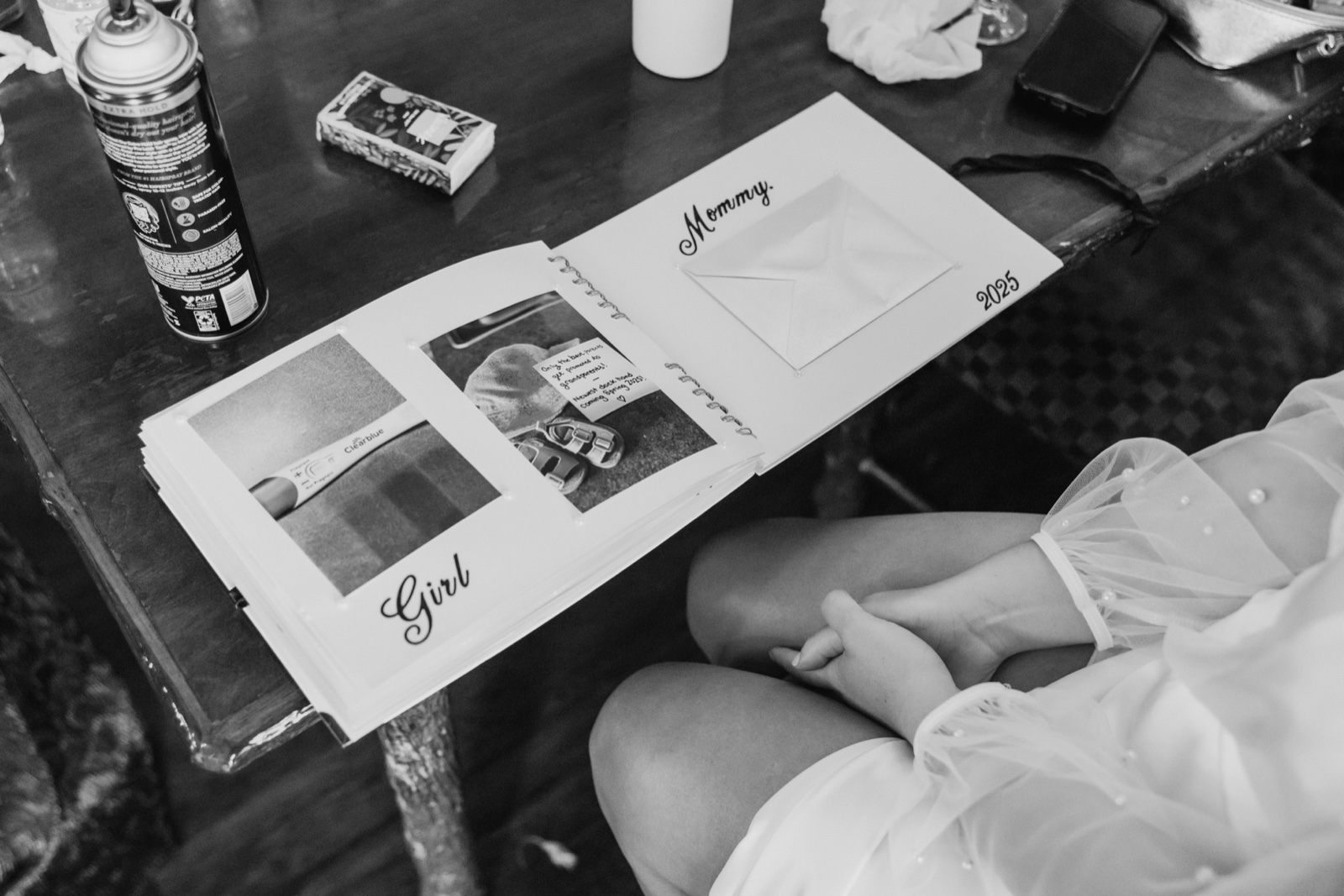 The bride in a pearl-trimmed veil holds an open scrapbook on her lap showing a positive pregnancy test, baby shoes, and a handwritten note announcing a baby girl coming spring — Tim Larsen Photography, Brainerd Lakes MN