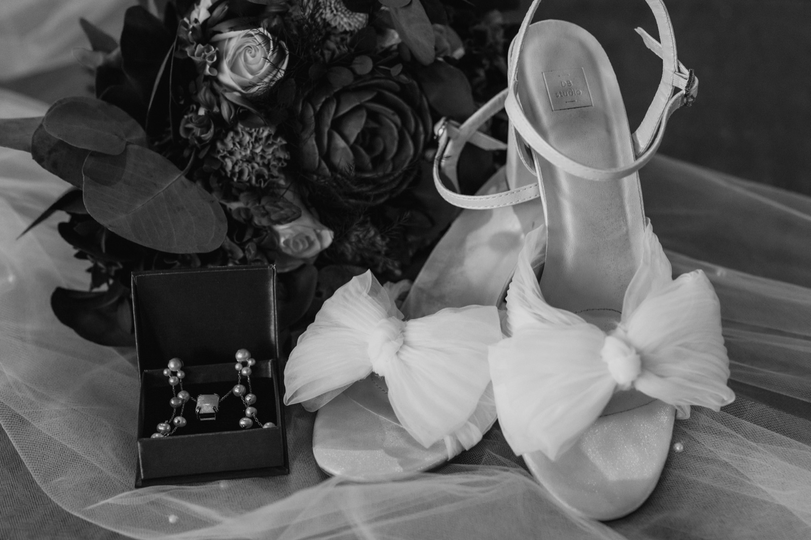 A black-and-white detail composition of the bride's bouquet, an open jewelry box with earrings and a pendant, and a pair of bow-adorned heels arranged on a veil — Tim Larsen Photography, Brainerd Lakes MN