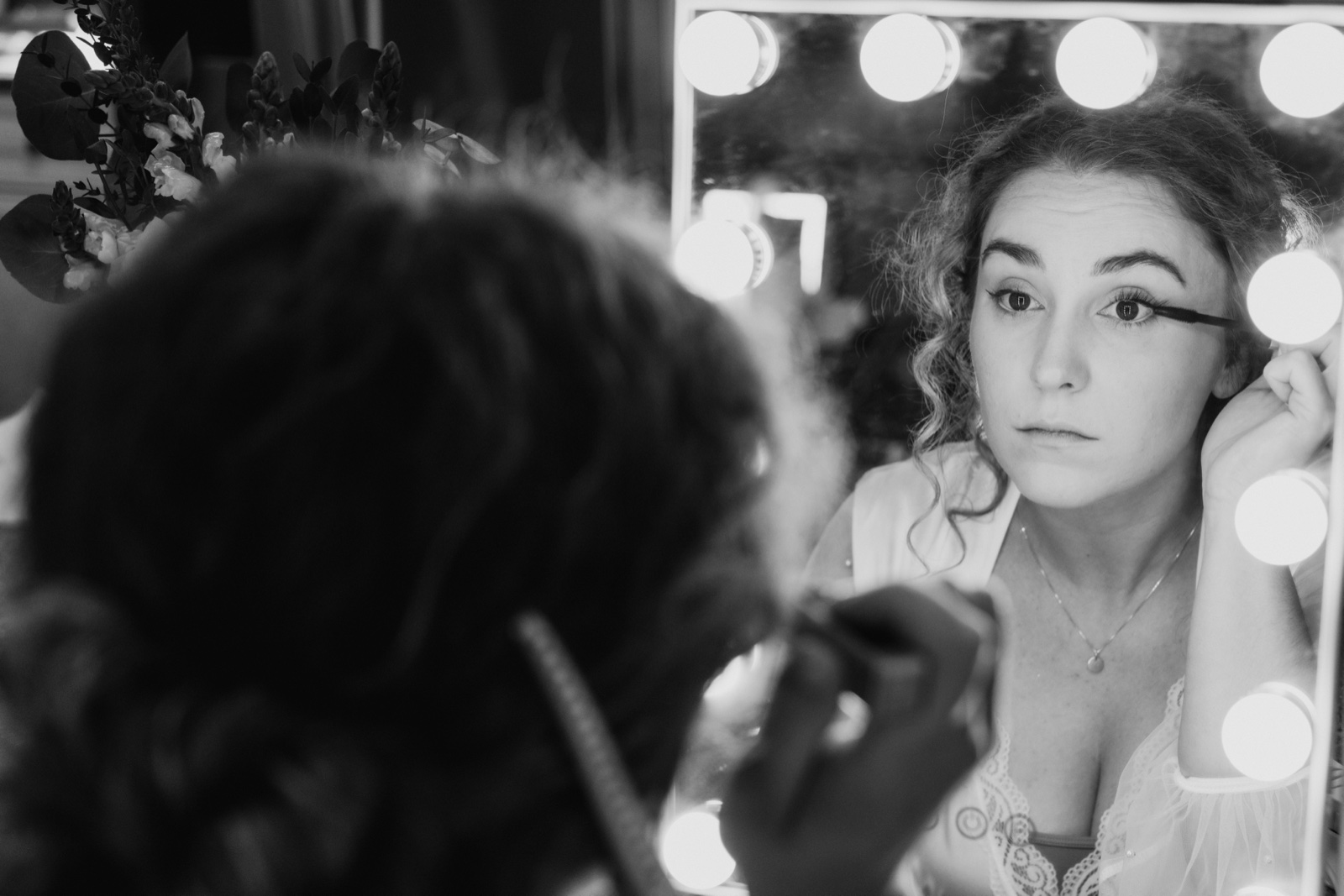 A black-and-white over-the-shoulder view of the bride applying mascara, her face reflected in a bulb-lit vanity mirror with florals to the side — Tim Larsen Photography, Brainerd Lakes MN