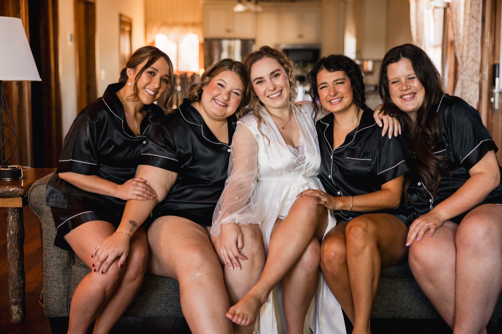 The bride in a sheer white robe sits on a couch flanked by four bridesmaids in matching black satin pajama sets, all smiling at the camera in a warmly lit cabin interior — Tim Larsen Photography, Brainerd Lakes MN