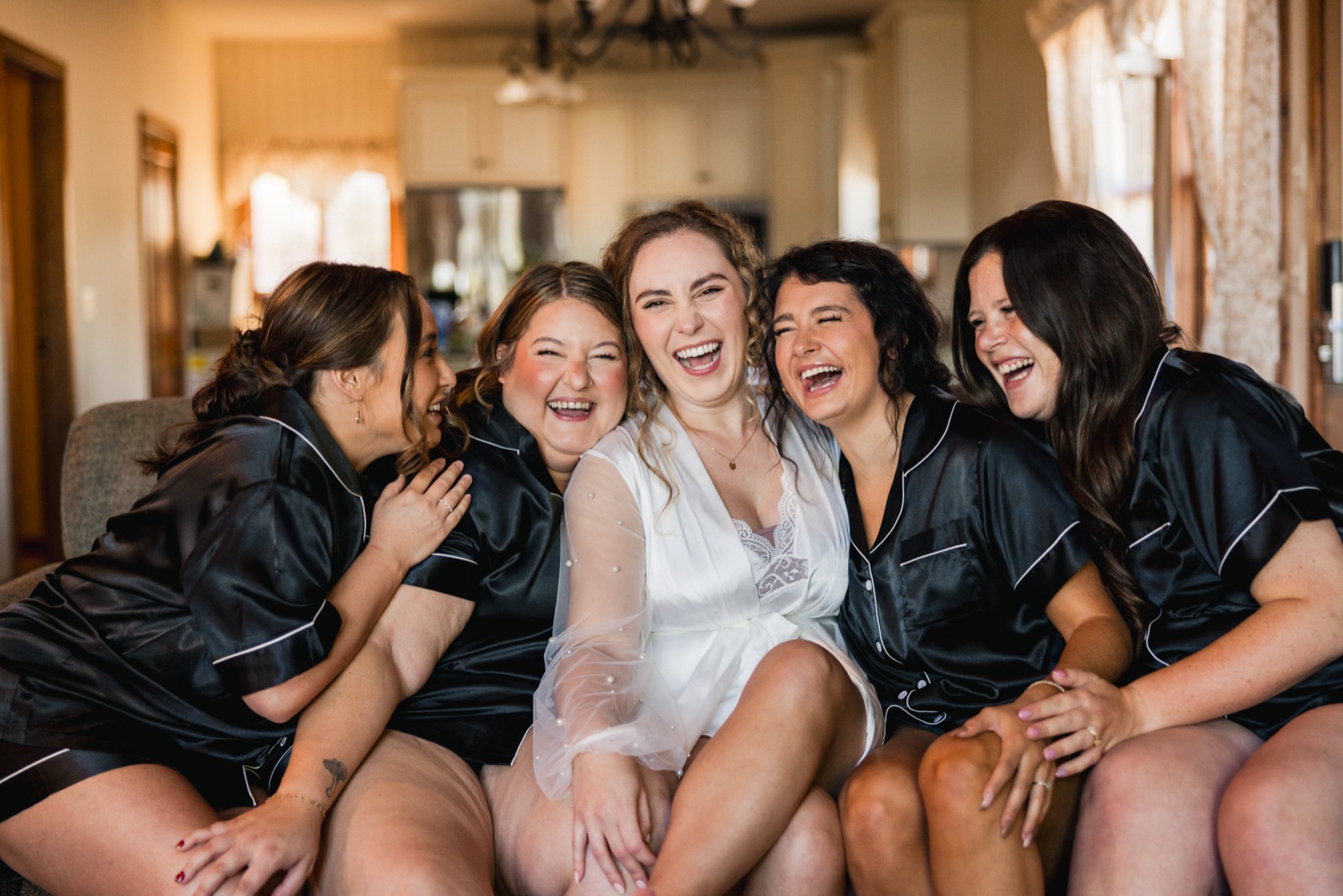 The bride sits on a couch in a sheer white robe surrounded by four bridesmaids in black satin pajamas, all laughing together in window light — Tim Larsen Photography, Brainerd Lakes MN