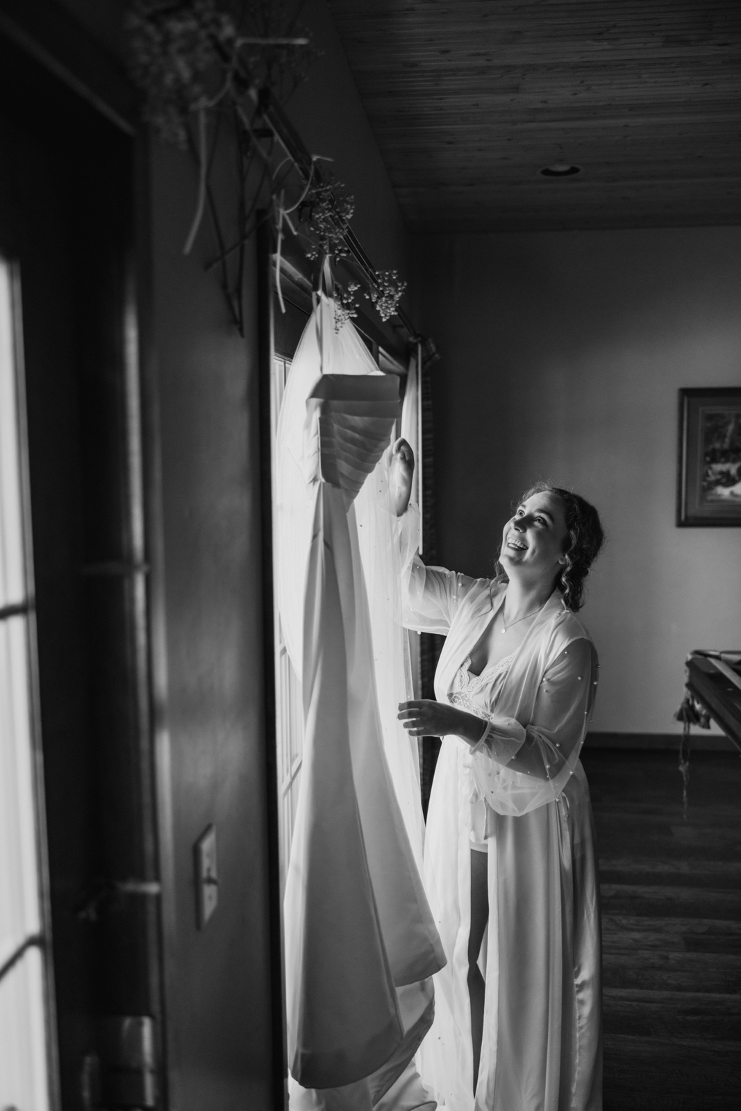 The bride in a silk robe reaches up toward her wedding dress hanging in a window, smiling and looking up at it — Tim Larsen Photography, Brainerd Lakes MN