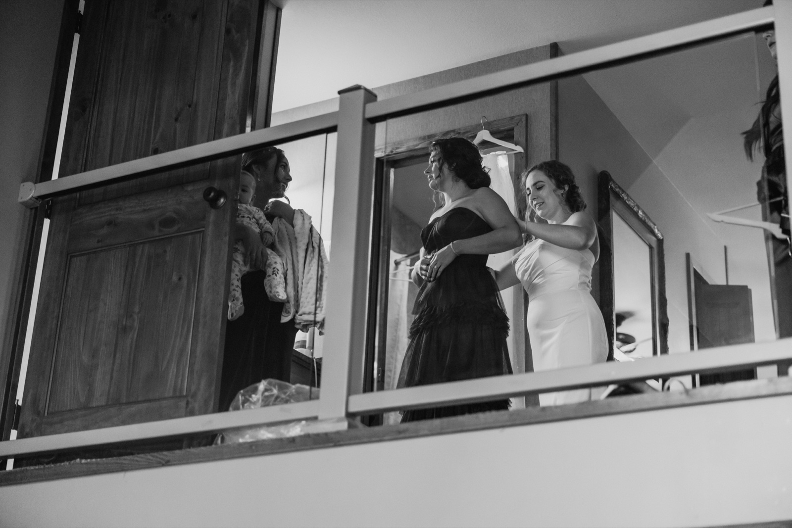A bridesmaid in a dark gown helps fasten the back of the bride's white dress on an upstairs landing, framed through wooden railing balusters with a mirror reflection visible at left — Tim Larsen Photography, Brainerd Lakes MN