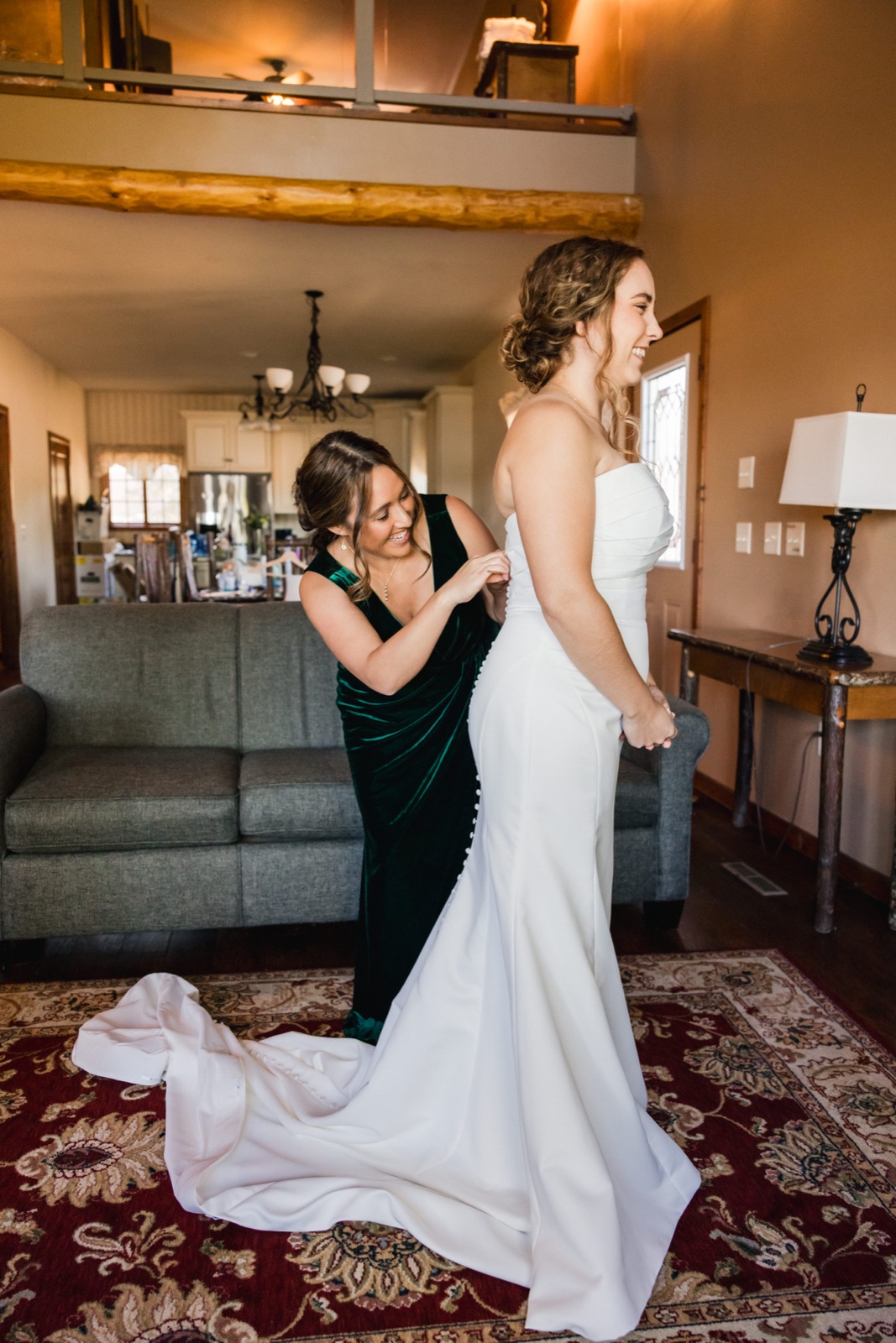 The bride laughs as a woman in an emerald velvet dress fastens the buttons on the back of her gown in a cabin living room with the train spilling onto a patterned rug — Tim Larsen Photography, Brainerd Lakes MN