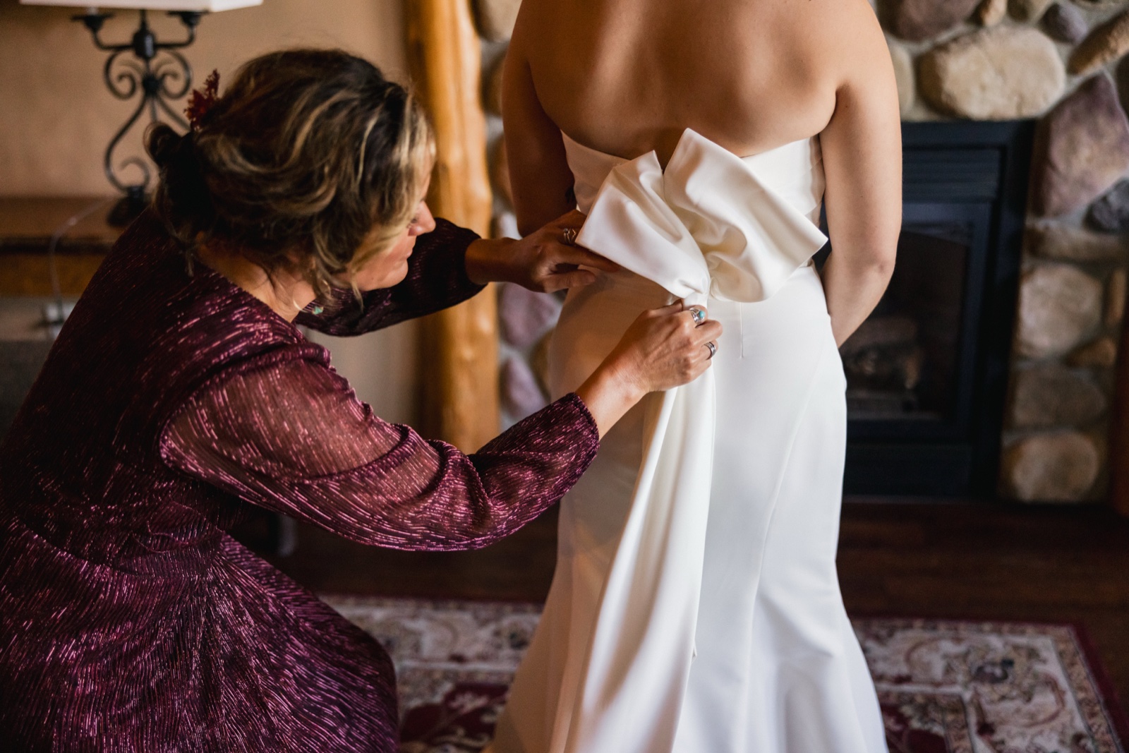 A woman in a burgundy dress kneels to fasten the back of the bride's white gown in a room with a stone fireplace — Tim Larsen Photography, Brainerd Lakes MN