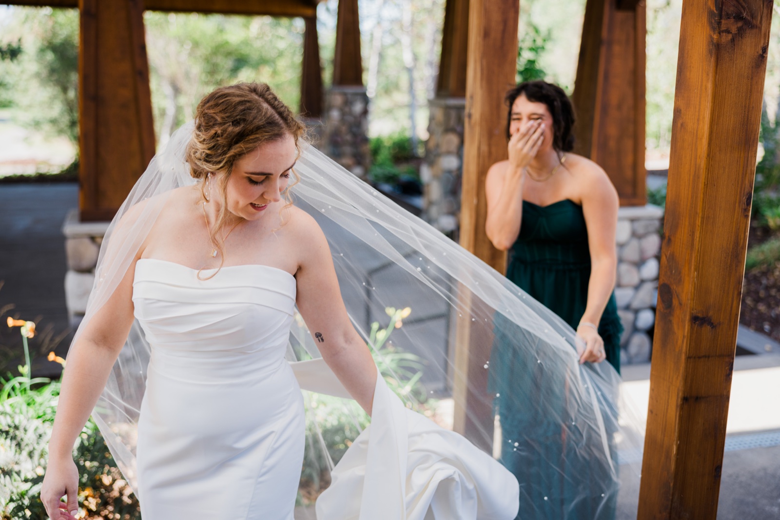 The bride walks beneath a wooden pavilion with her veil trailing behind her as a bridesmaid in a green dress covers her mouth, visibly emotional in the background — Tim Larsen Photography, Brainerd Lakes MN